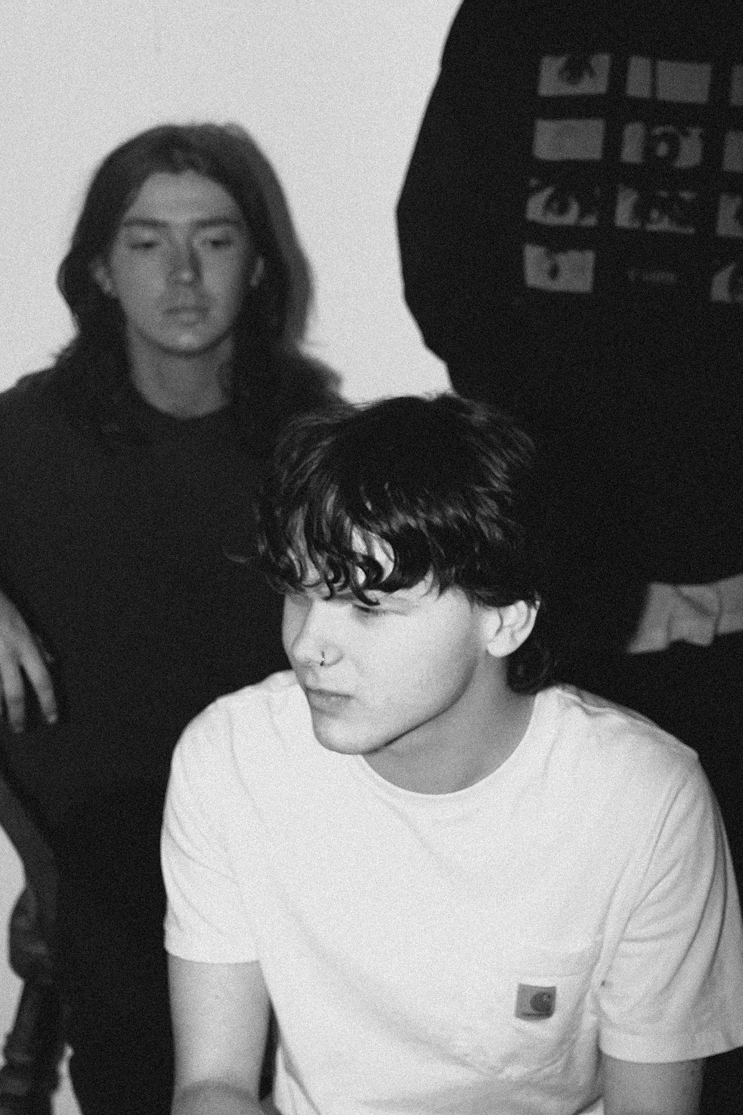 Black and white photo of two young men, one with curly hair and a nose piercing, sitting and looking downward, the other with long straight hair looking straight ahead, with a person holding a phone in the background.