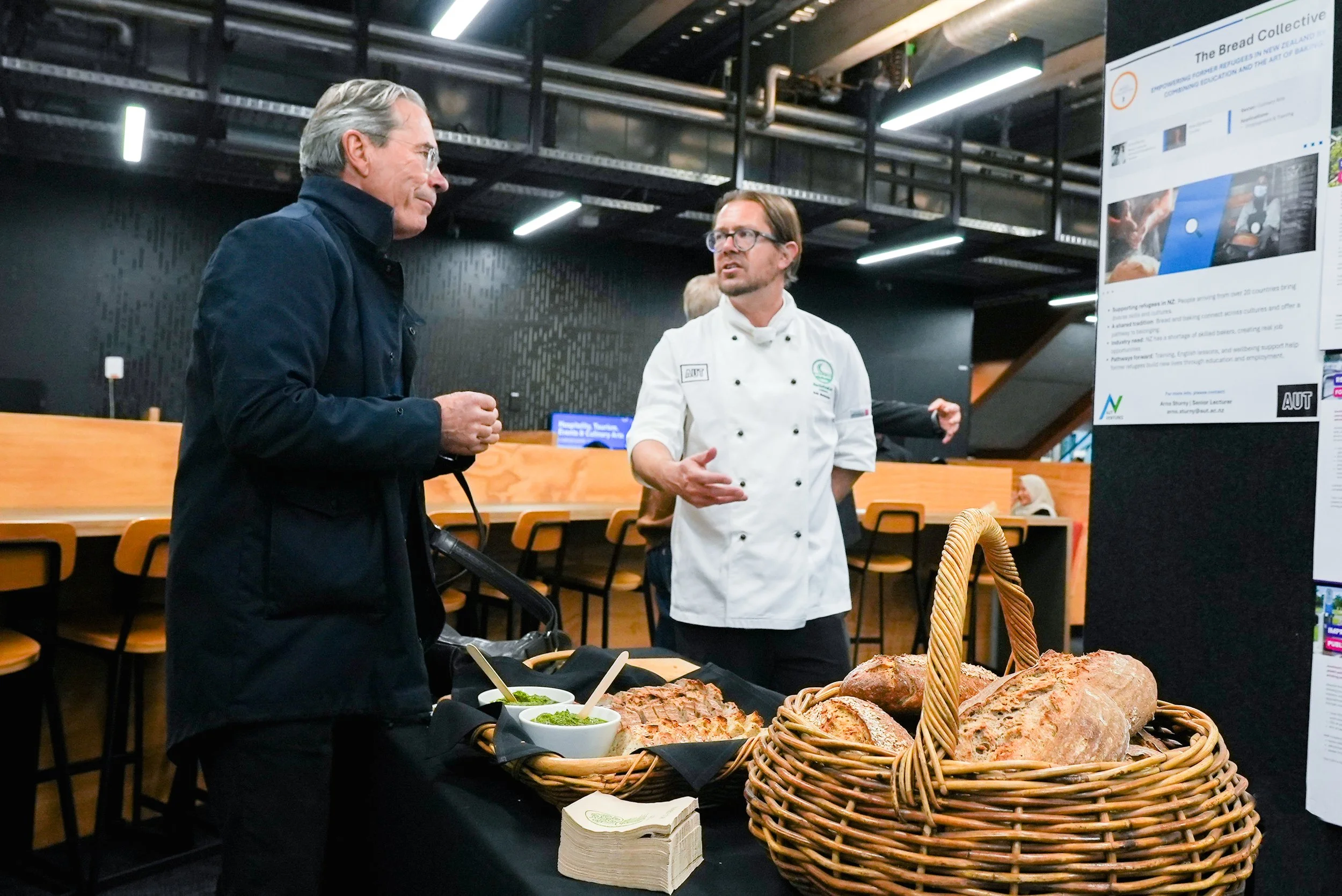 Two men engaged in conversation at a food display table with bread and bowls of green spread, in a modern indoor setting.