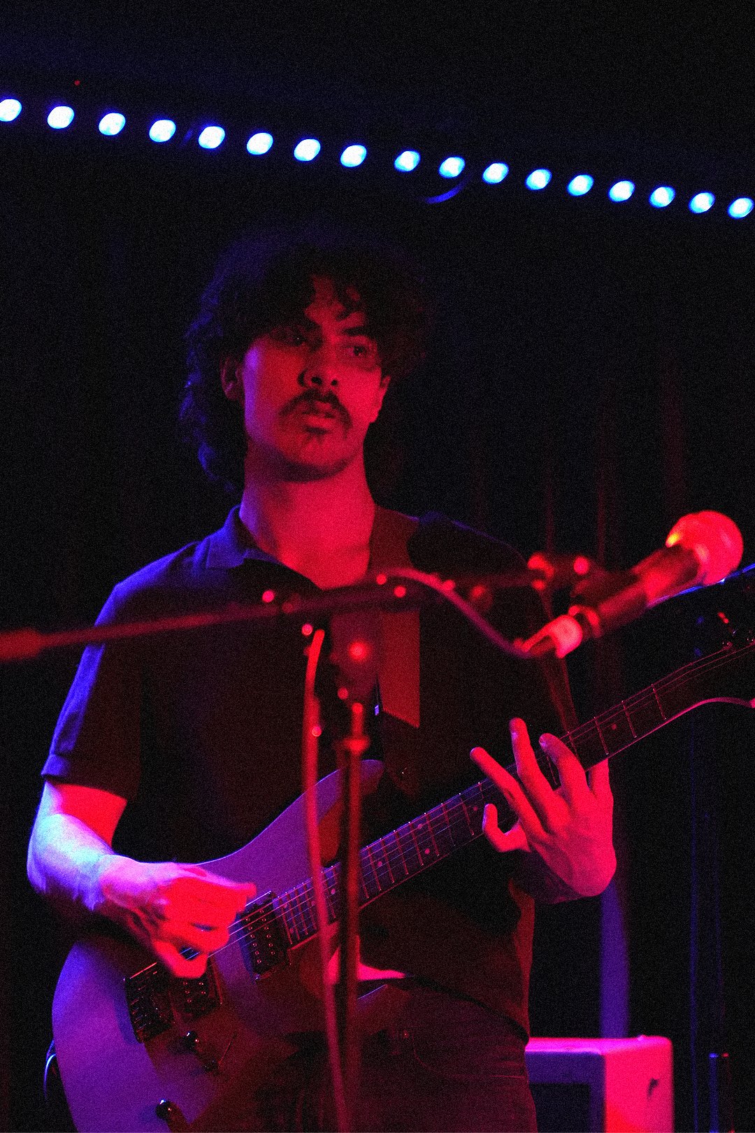 A man with dark curly hair and a mustache plays an electric guitar on stage, illuminated by red and purple stage lights, with a microphone in front of him.