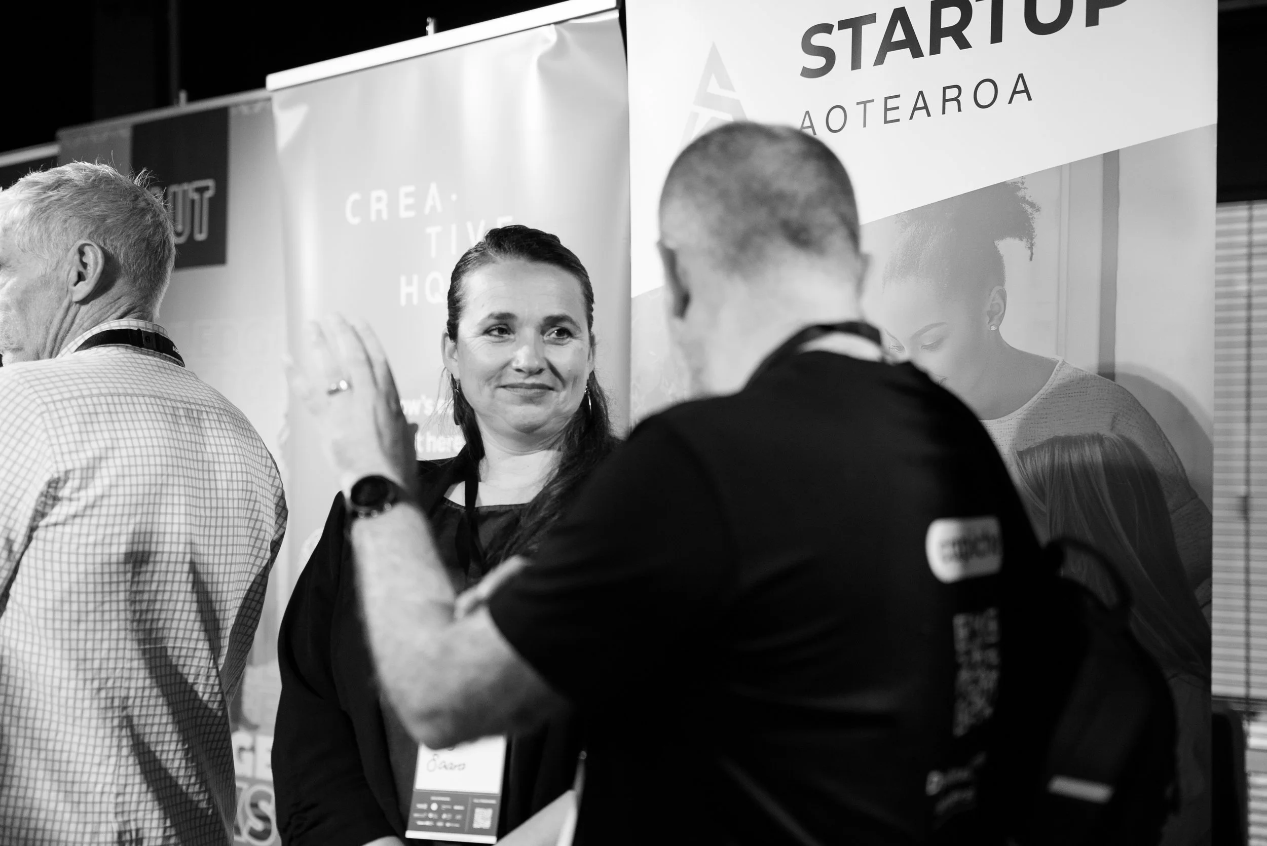 Three people are engaged in conversation at a startup event, with one woman smiling and talking to a man, while another man faces away. Background displays banners labeled 'STARTUP' and 'CREATIVE'.