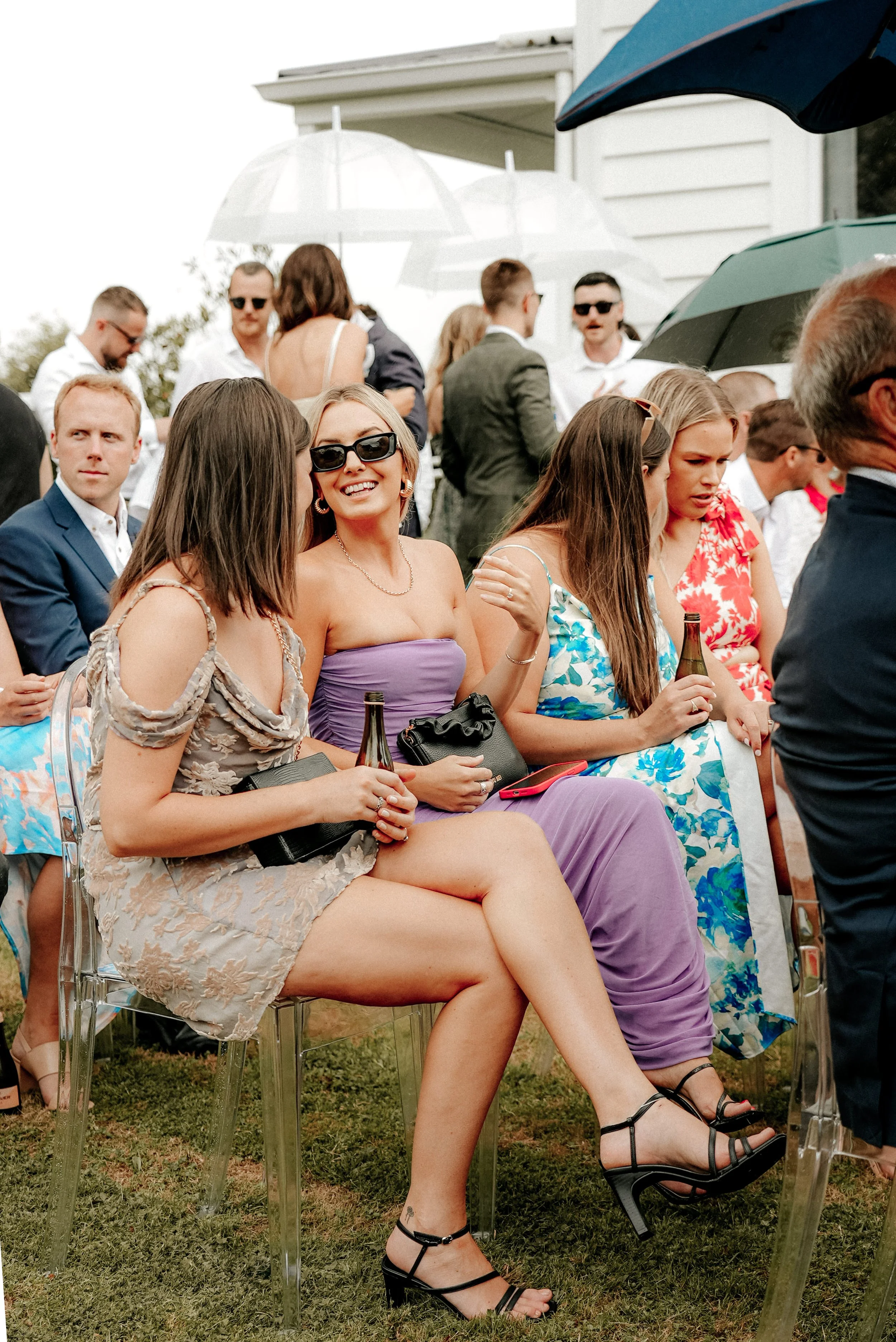 People gathered outdoors at an event, some holding umbrellas, with a group seated in the foreground including women in colorful dresses, one wearing sunglasses and smiling.