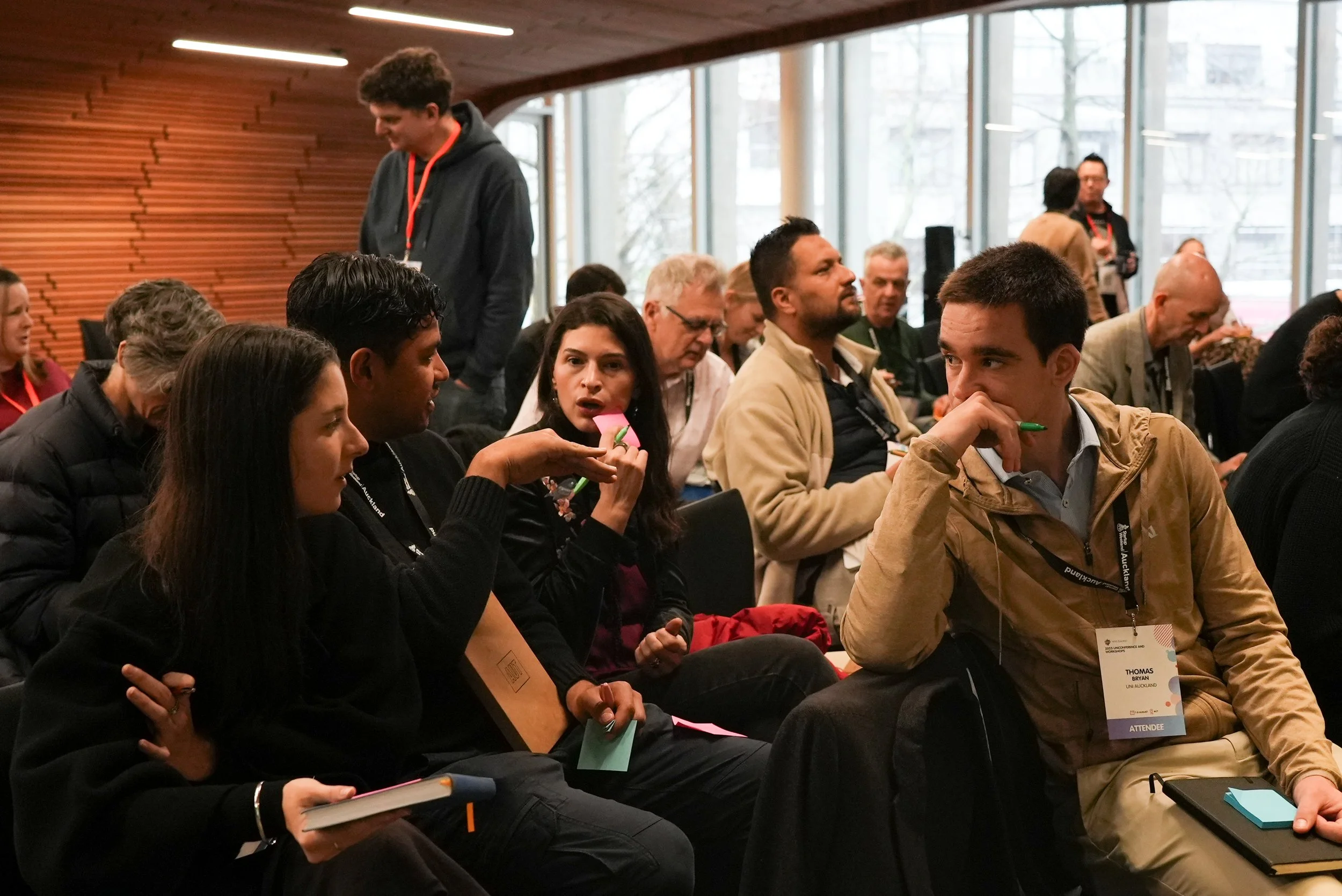 Group of people attending a conference or seminar, sitting and engaging in conversation, with some taking notes or listening attentively.