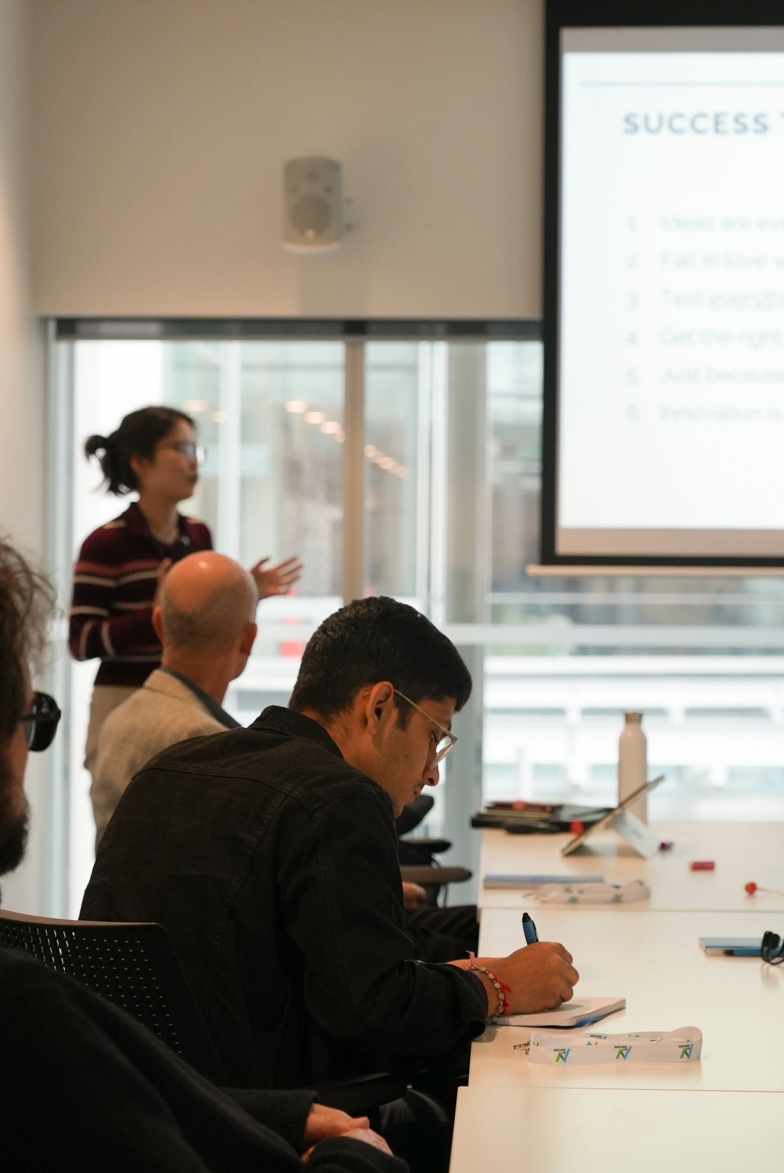 A woman giving a presentation in front of a projection screen with a slide titled 'SUCCESS'. Several people are seated at a table, listening, taking notes, and looking at the screen.