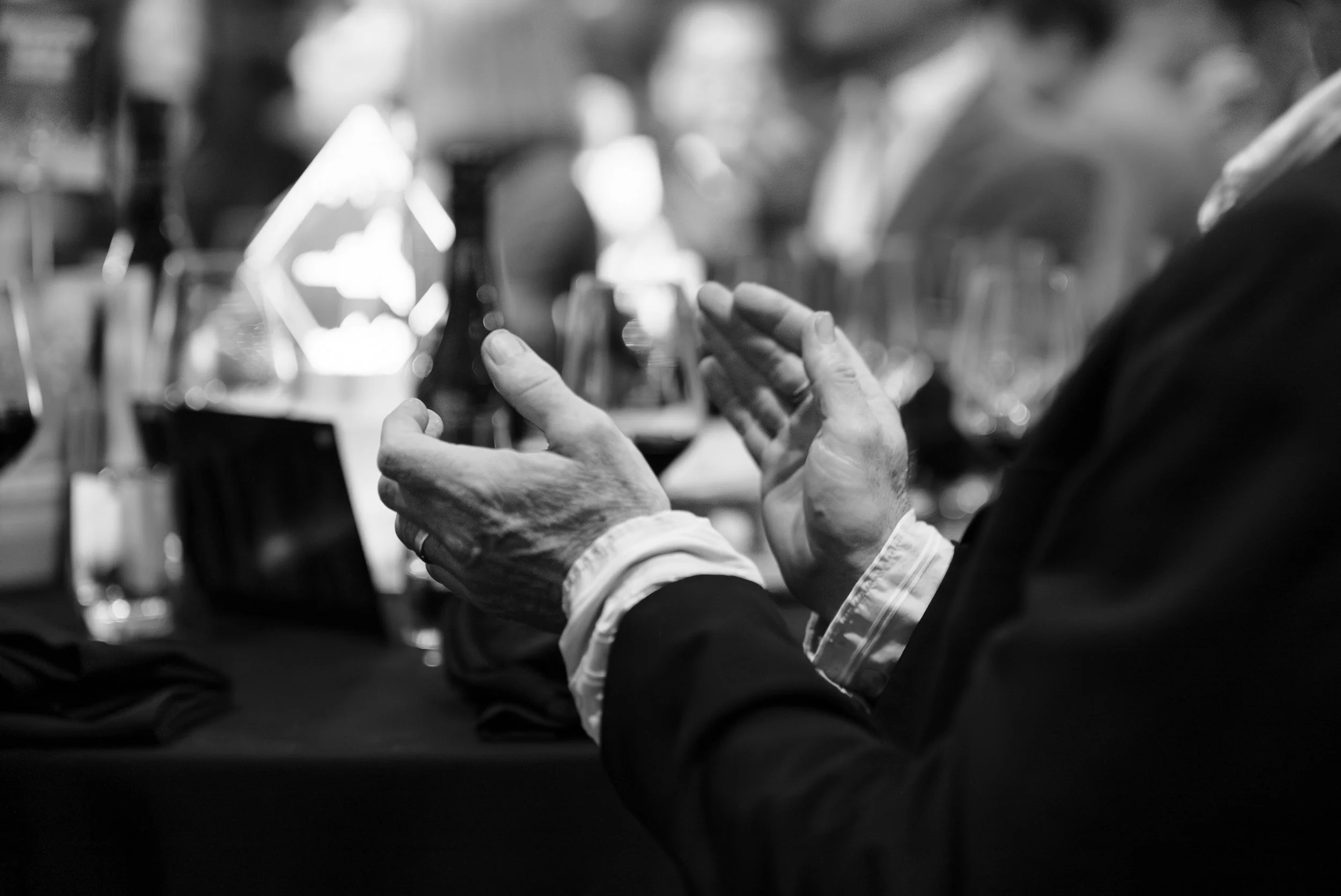 Close-up of hands clapping at a formal event, with a blurred background of glasses and other people.