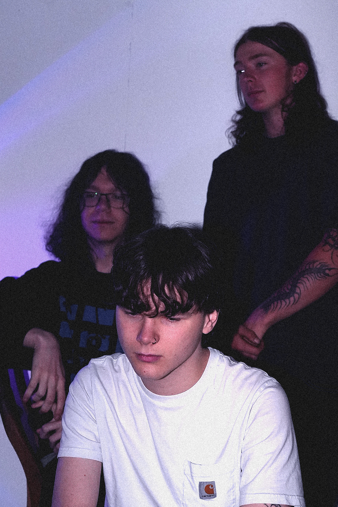 Three young adults, two women and one man, in a dimly lit room with purple lighting. The man in the foreground has black, curly hair, a nose ring, and is wearing a white Carhartt t-shirt. The woman on the left has shoulder-length dark hair, glasses, and a black hoodie. The woman on the right has long curly hair, tattoos on her arm, and is wearing a black shirt.