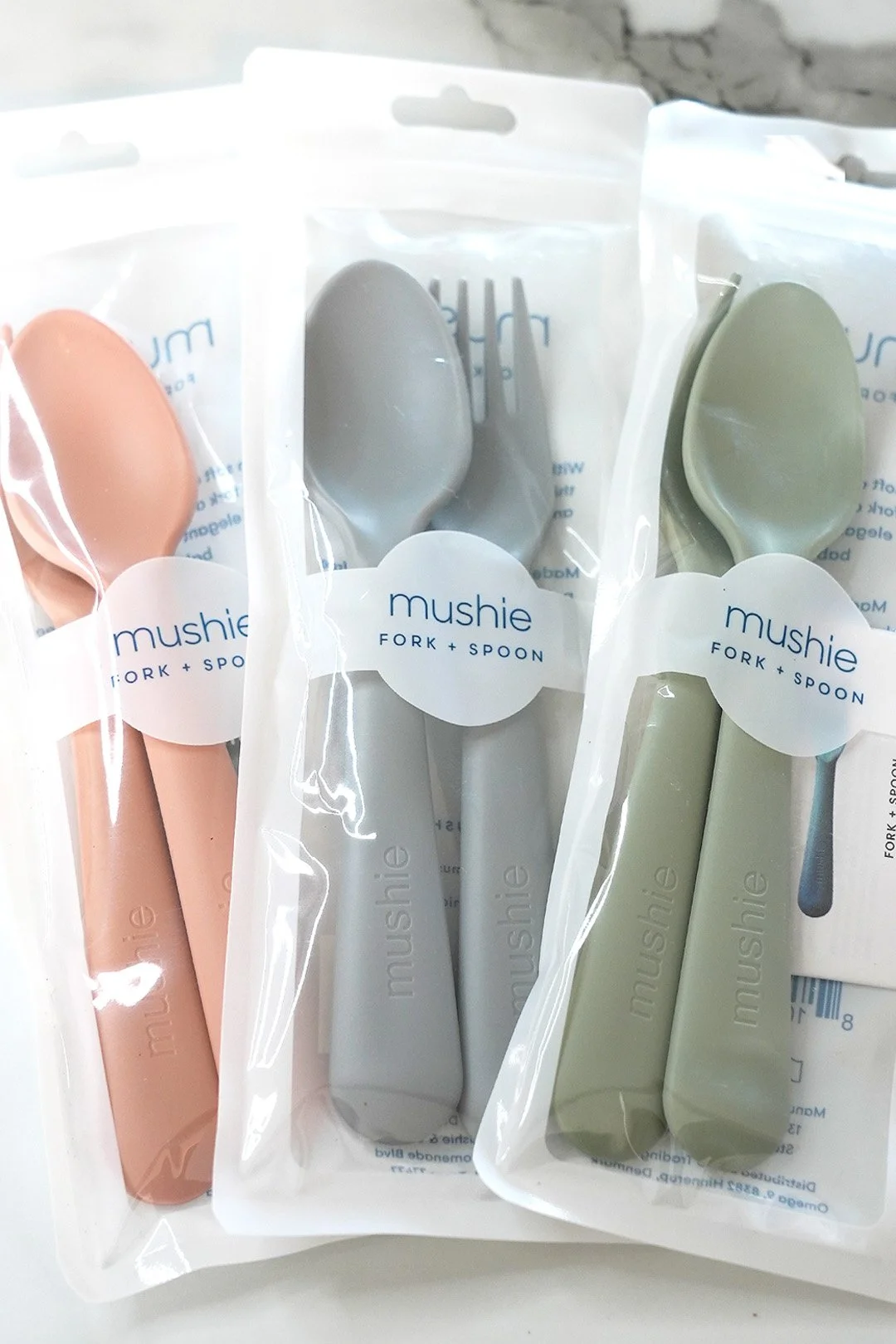 Sets of pastel-colored baby utensils including soft pink, grey, and olive green, each in individual clear plastic packaging labeled 'mushie fork + spoon'.