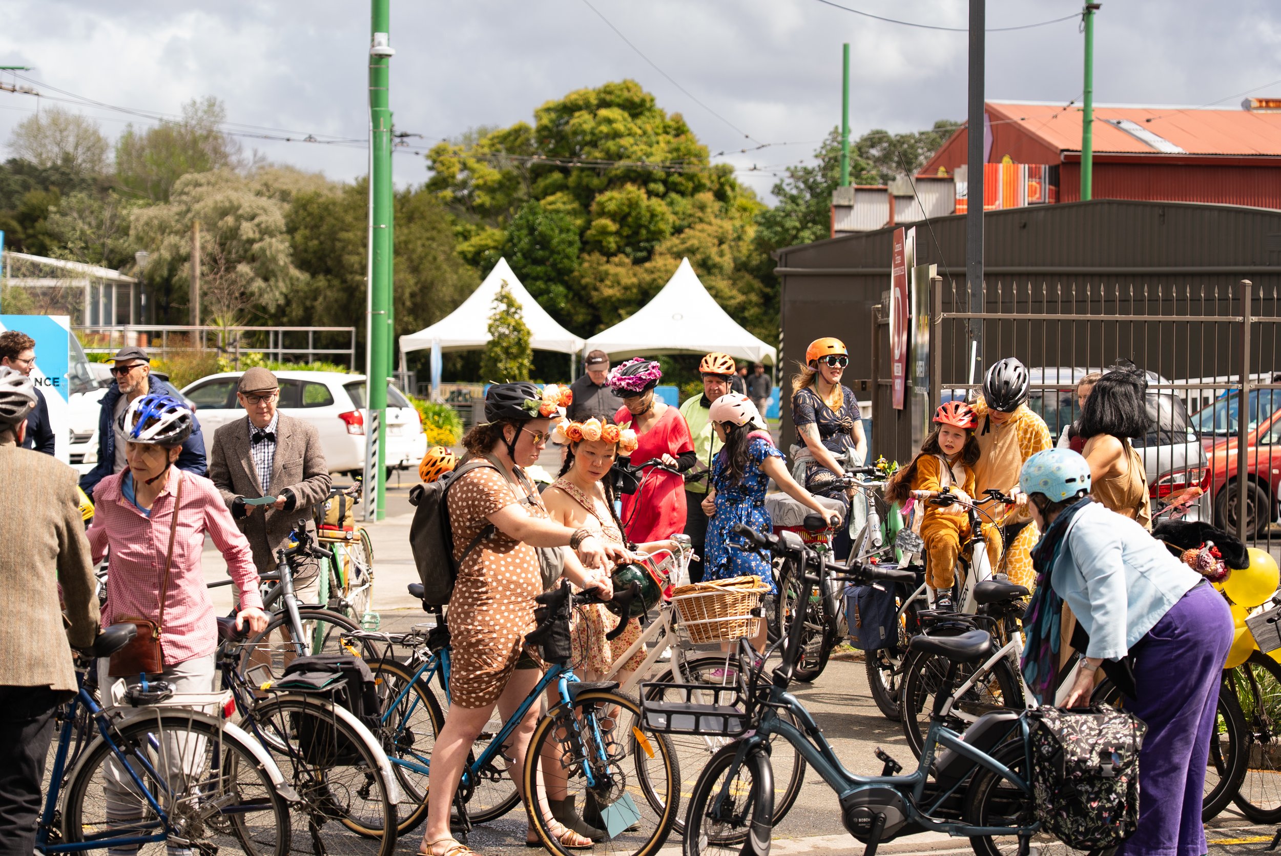 Group of people with bicycles, some wearing helmets and costumes, outdoors near tents and cars, on a cloudy day.
