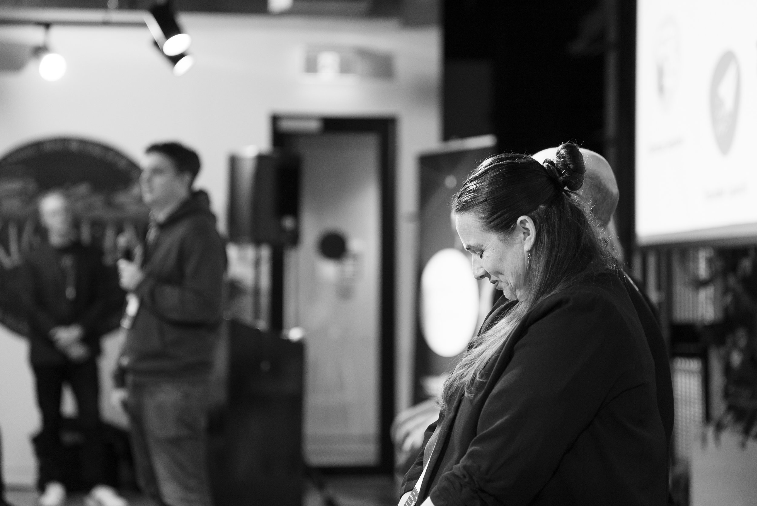 Black and white photo of woman with long hair tied back, standing and looking down in a conference or presentation setting, with people in the background.