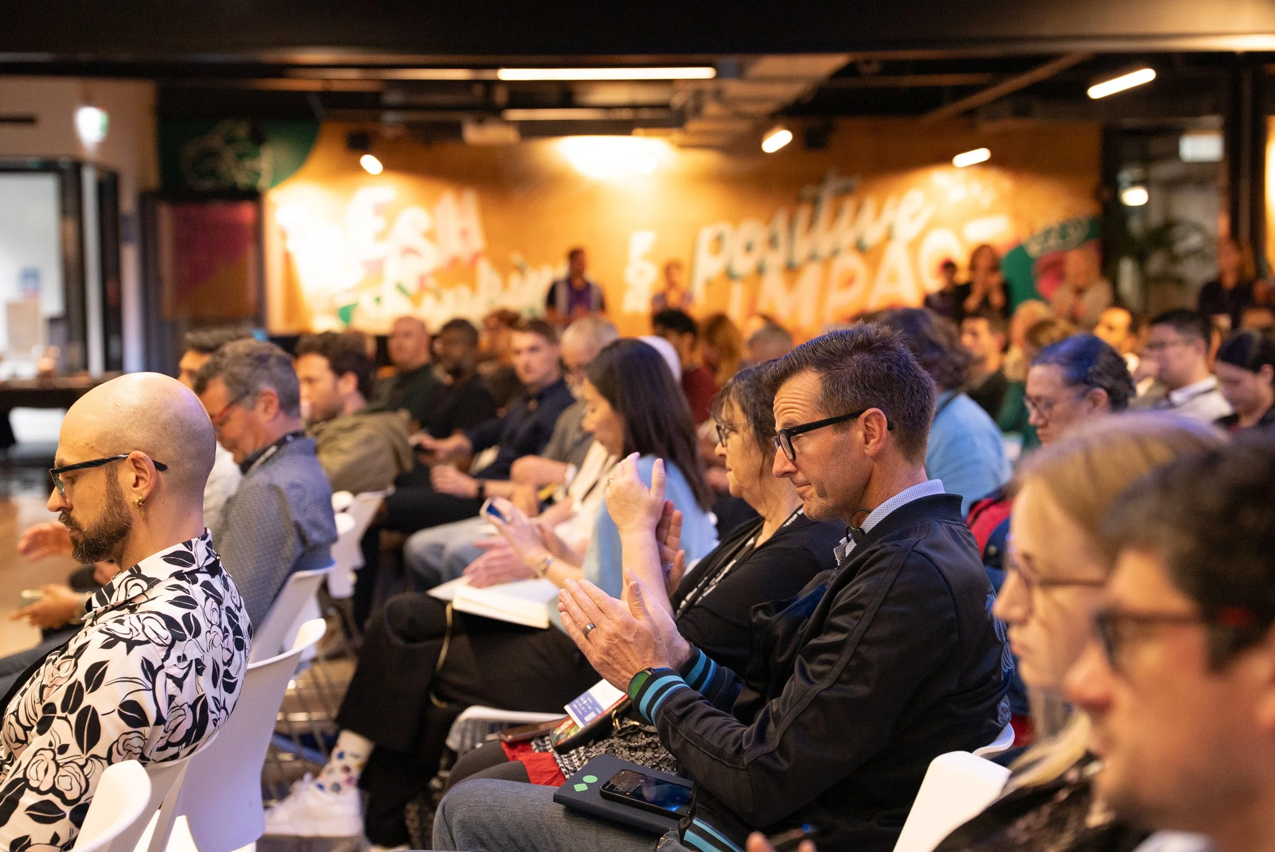 Attendees seated in rows at a conference or seminar, some looking at their phones, with a colorful mural background reading 'Esa' and 'Poblano'.