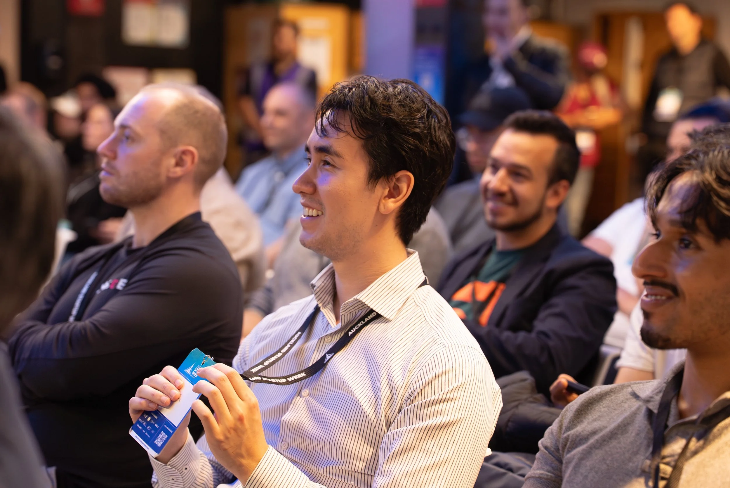 A group of men sitting and smiling at an indoor event or conference, with some wearing conference badges around their necks.