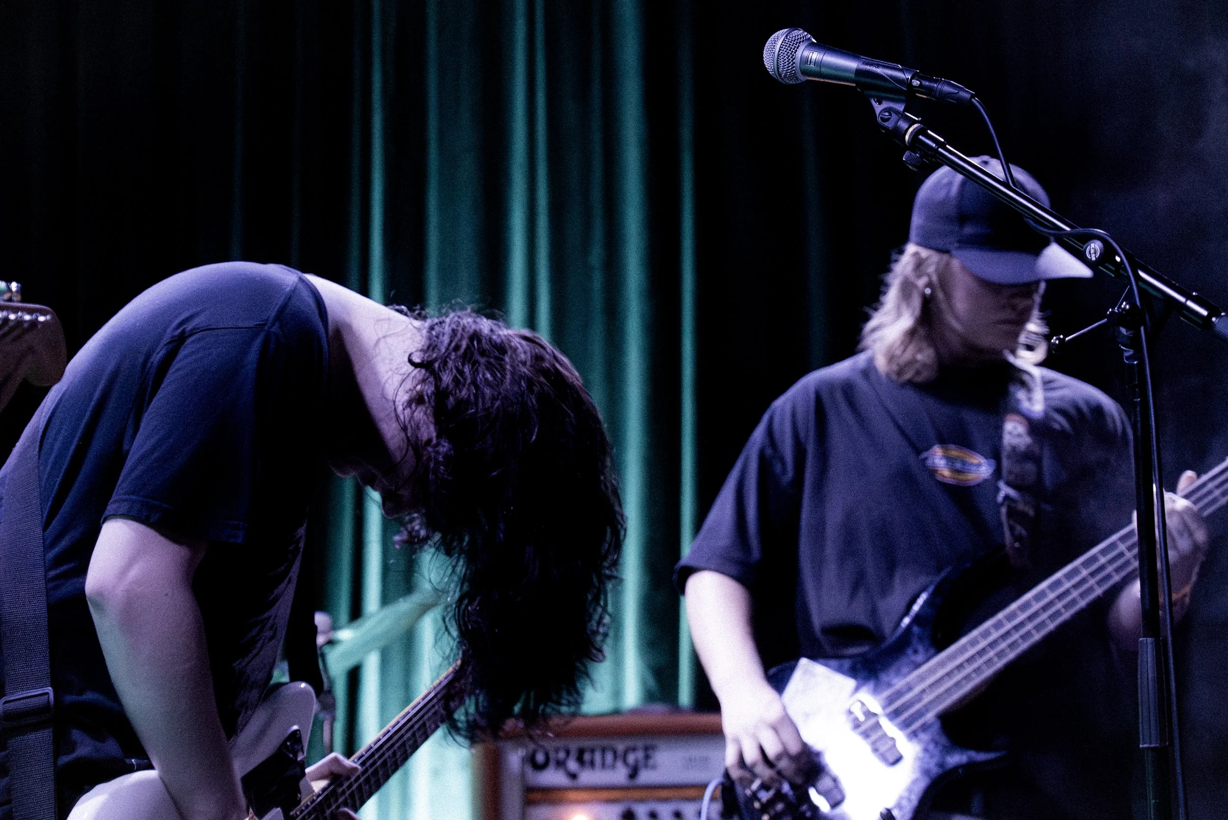 Two musicians performing on stage; one is playing electric guitar, and the other is playing bass guitar, with a microphone setup nearby. The stage has dark and green curtains in the background.