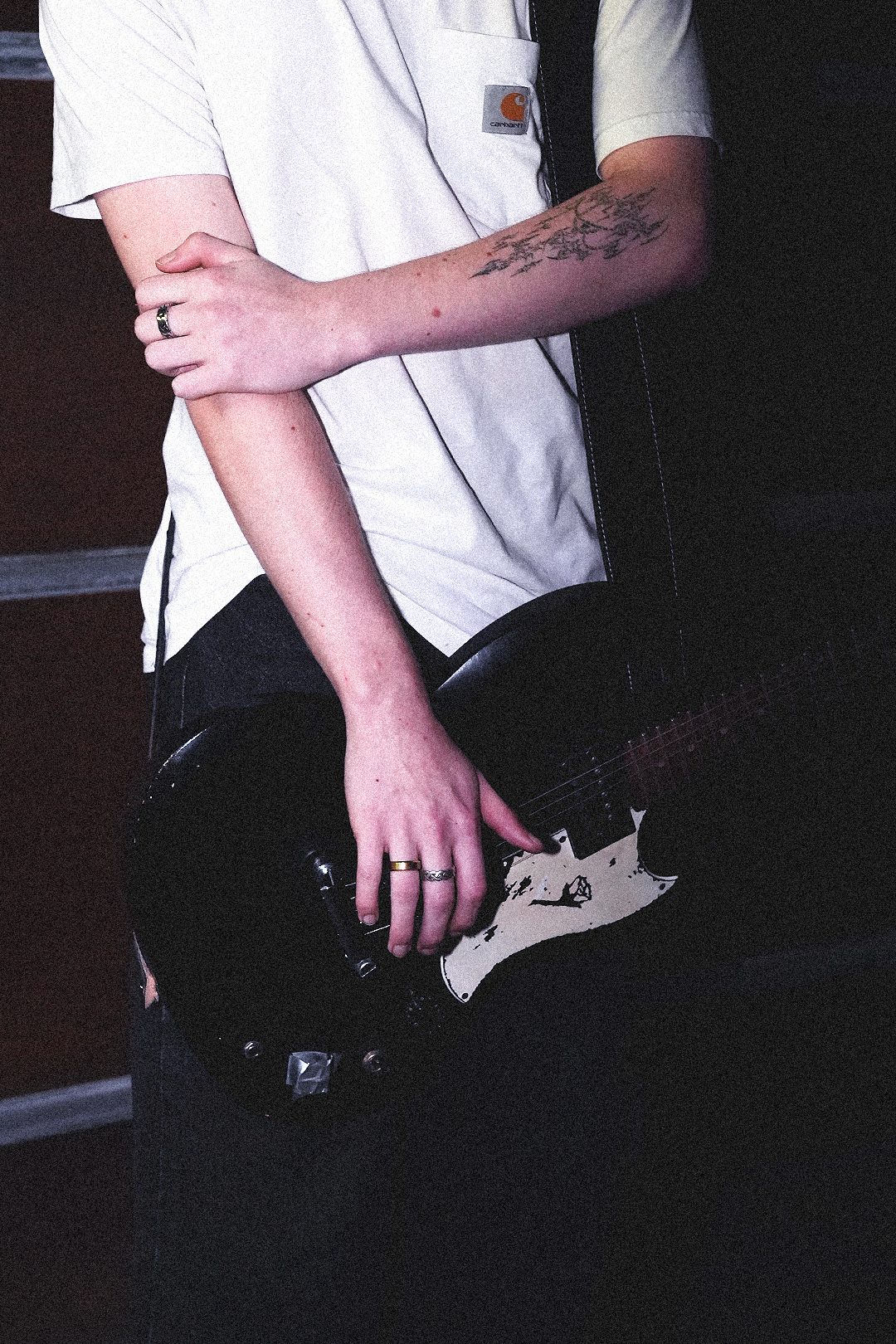 Person wearing a white Carhartt t-shirt, sitting on a black chair, holding a black electric guitar with a white pickguard, with tattoos on their right arm and rings on their fingers.