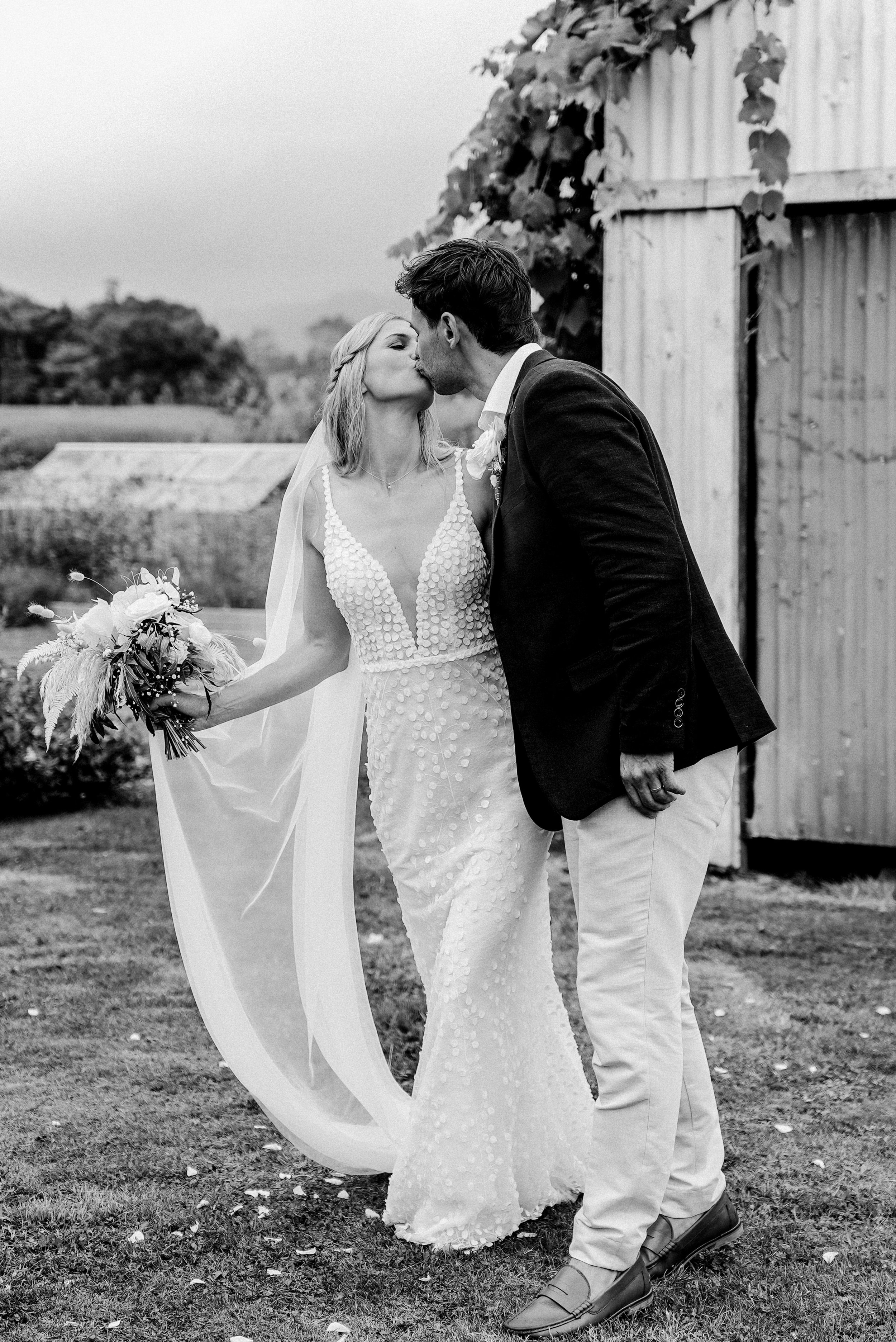 A bride and groom kissing outdoors during their wedding, with the bride holding a bouquet, in black and white photo.