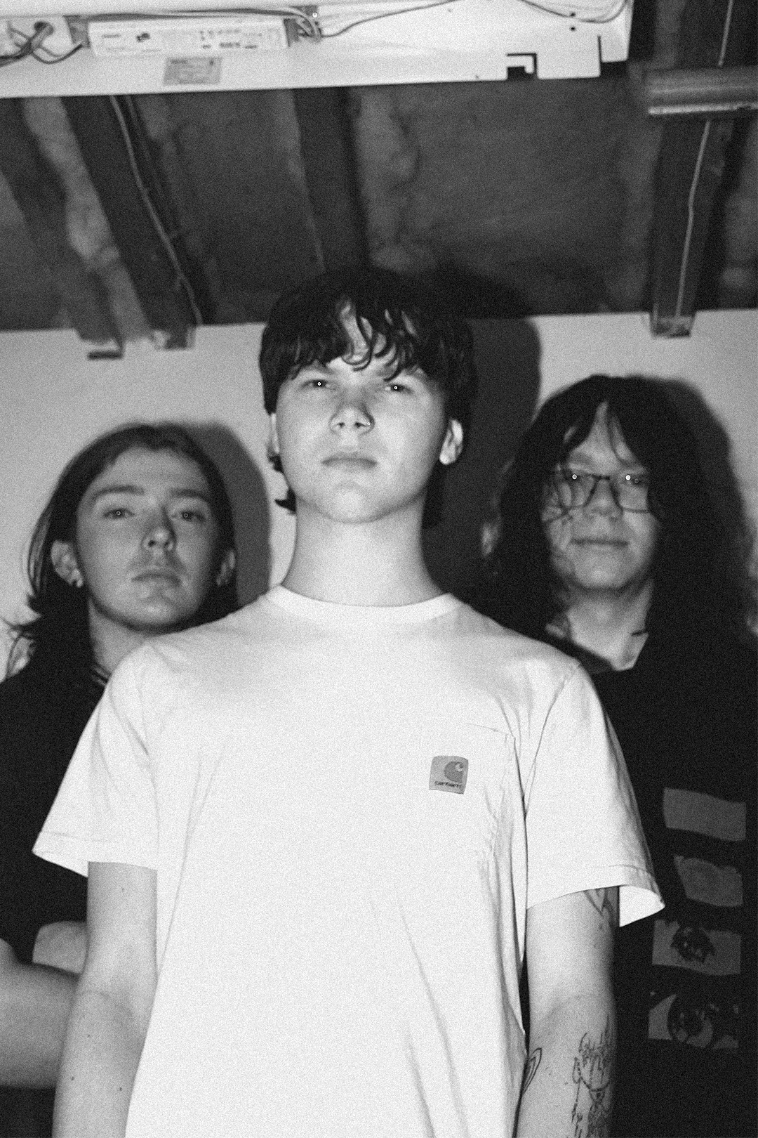Black and white photo of three young men standing indoors, with the person in the middle in the foreground. The person in the middle has short messy hair and is wearing a white T-shirt with a small logo on the chest. The person on the left has long straight hair, and the person on the right has long wavy hair and glasses. The background shows a ceiling with exposed wooden beams and some electrical wires.