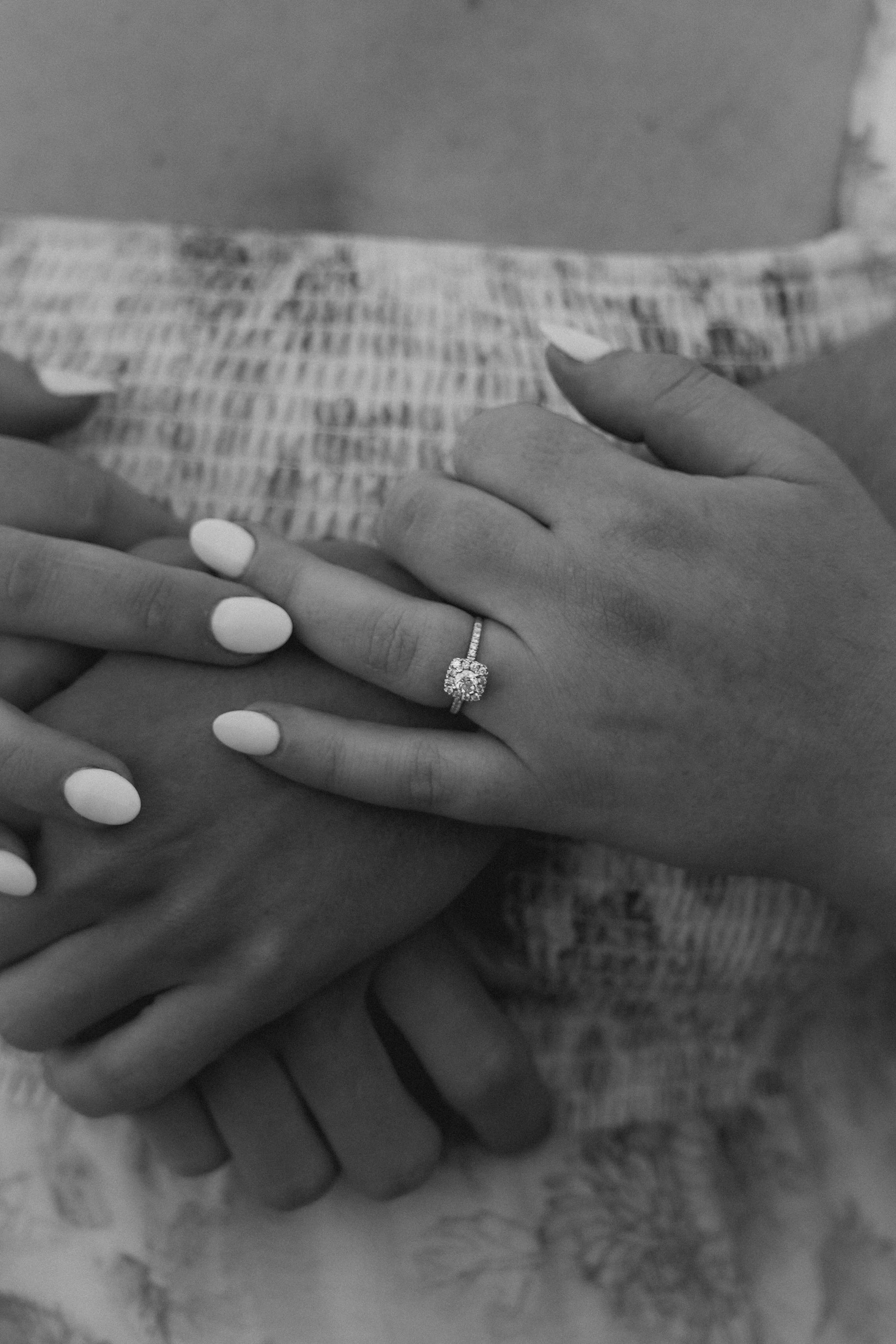 Close-up of two hands with wedding rings, one hand resting on top of the other, show a woman with white nail polish and an engagement ring with a large center diamond surrounded by smaller diamonds, on her ring finger, while the other hand has a ringless finger, all on a patterned fabric background.