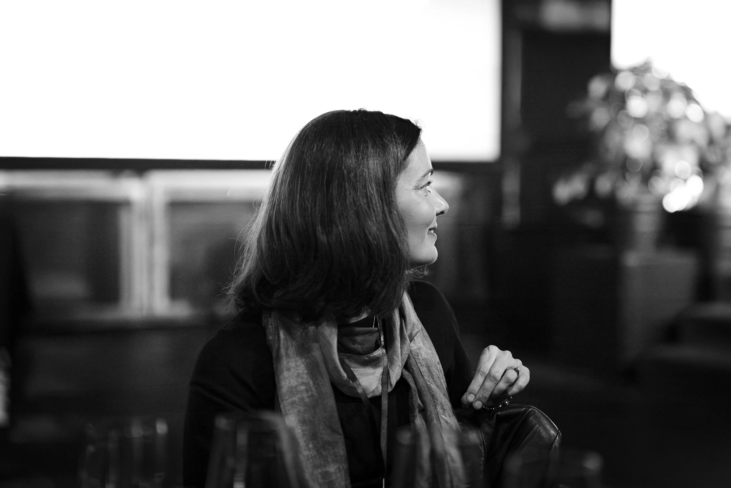 A woman with dark, shoulder-length hair sitting in a dimly lit restaurant or bar, smiling and looking to her right.