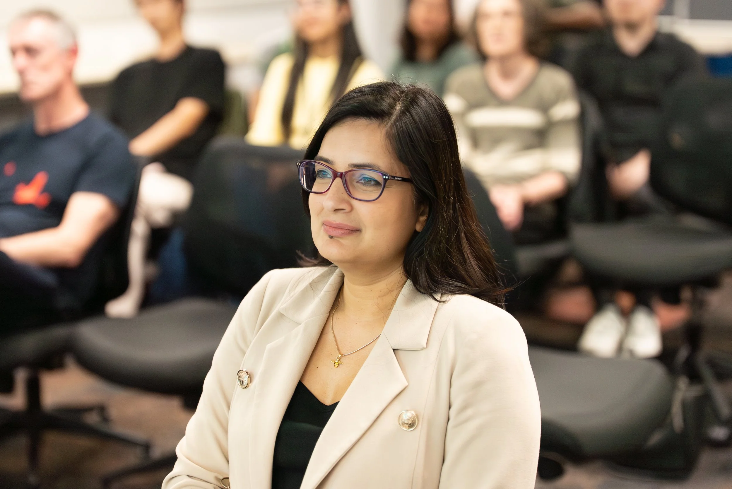 A woman with glasses and dark hair attending an indoor event, seated among others in a conference or seminar setting.