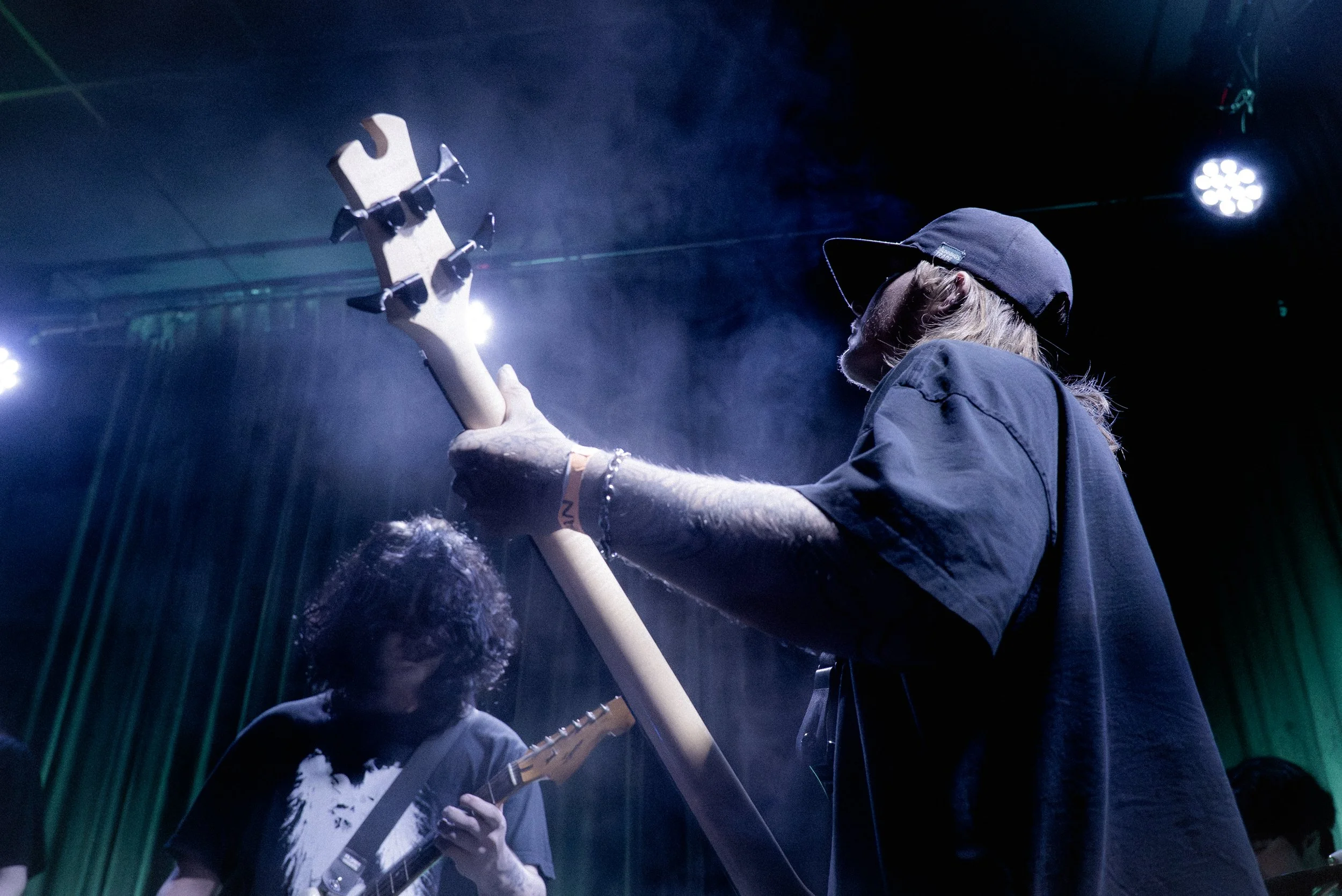 Two musicians performing on stage, one playing a guitar and the other playing a bass, with stage lights and green curtains in the background.