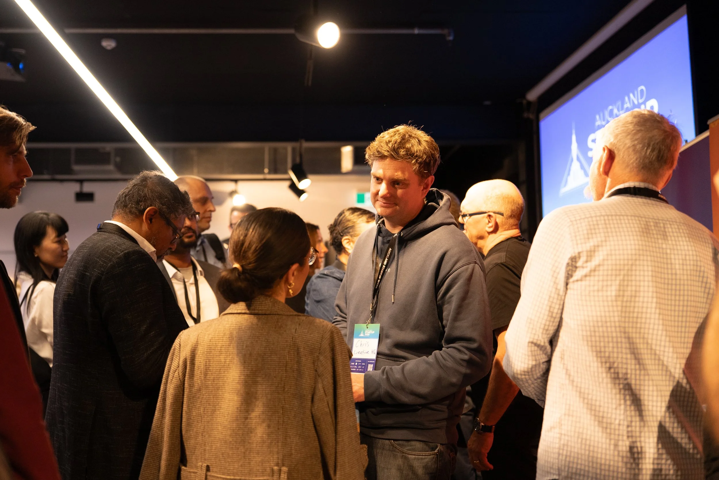 A group of diverse people at a conference or networking event, engaging in conversation, with a screen displaying 'AUCKLAND' in the background.
