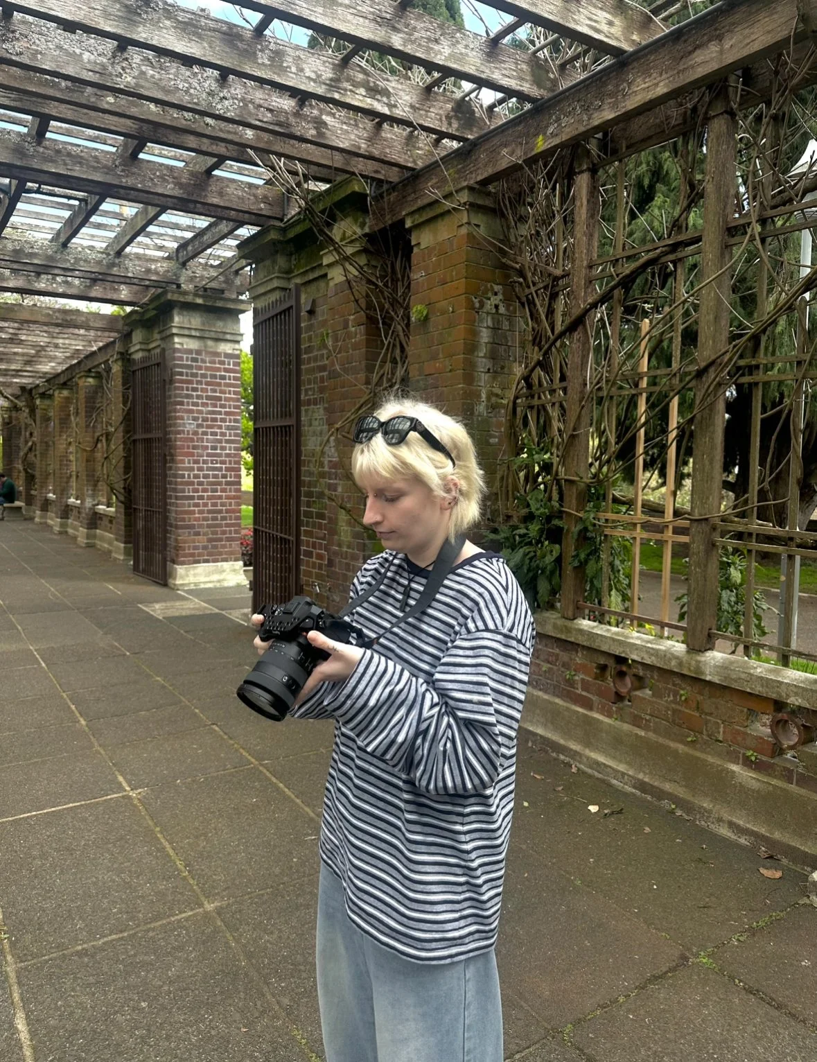 Young person with blond hair, wearing sunglasses on their head, looking at a camera they are holding, standing outdoors near brick pillars and a wooden trellis with vines.