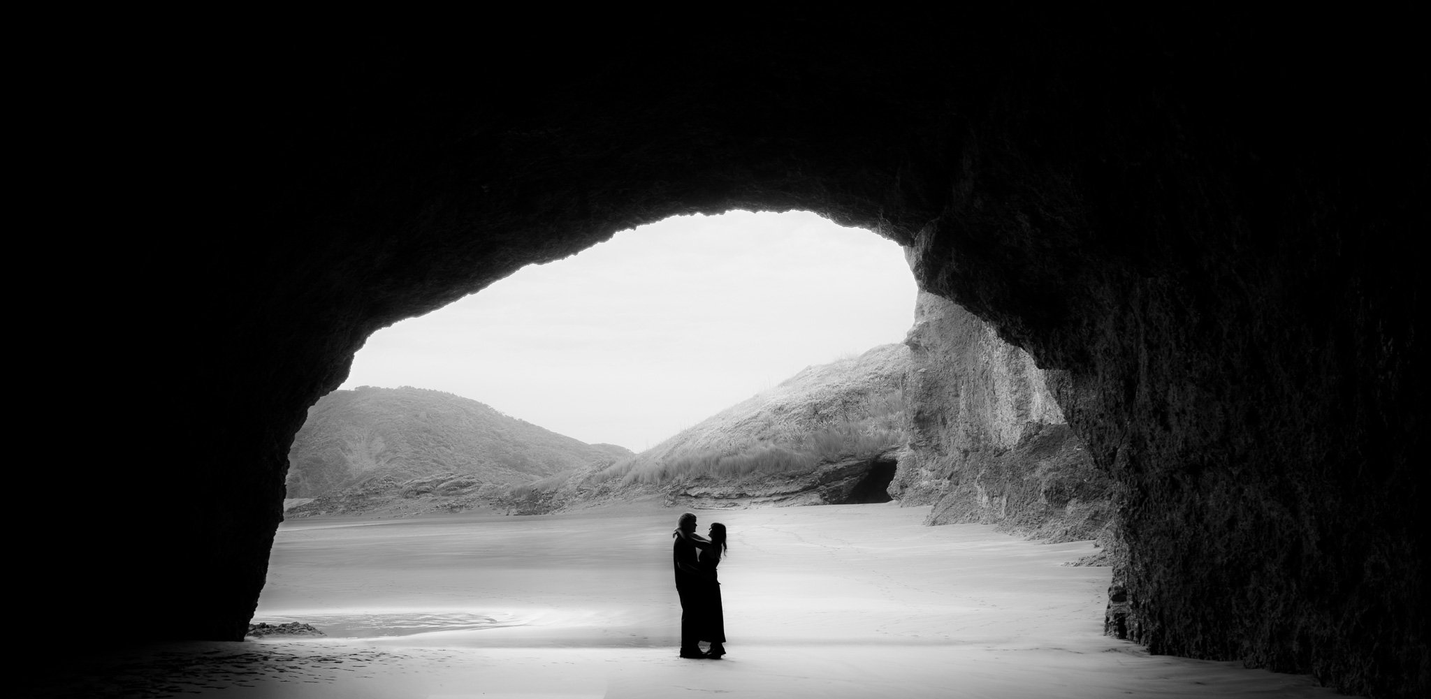 Couple embracing on the beach inside a large cave, with rocky hills and a cloudy sky in the background, in black and white