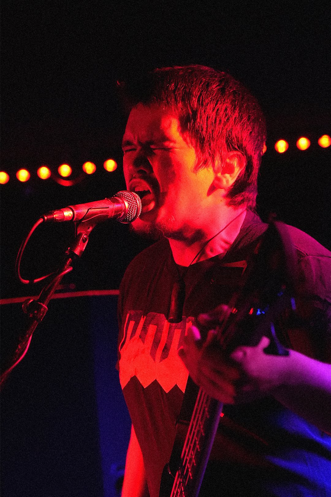 A man passionately singing into a microphone while playing an electric guitar in a dimly lit setting, illuminated by red and purple stage lights.