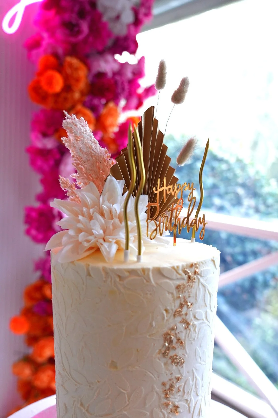 A tall white birthday cake decorated with pink and white flowers, golden accents, and a "Happy Birthday" topper. The background features a colorful flower arrangement with pink, orange, and purple flowers and a window with trees outside.