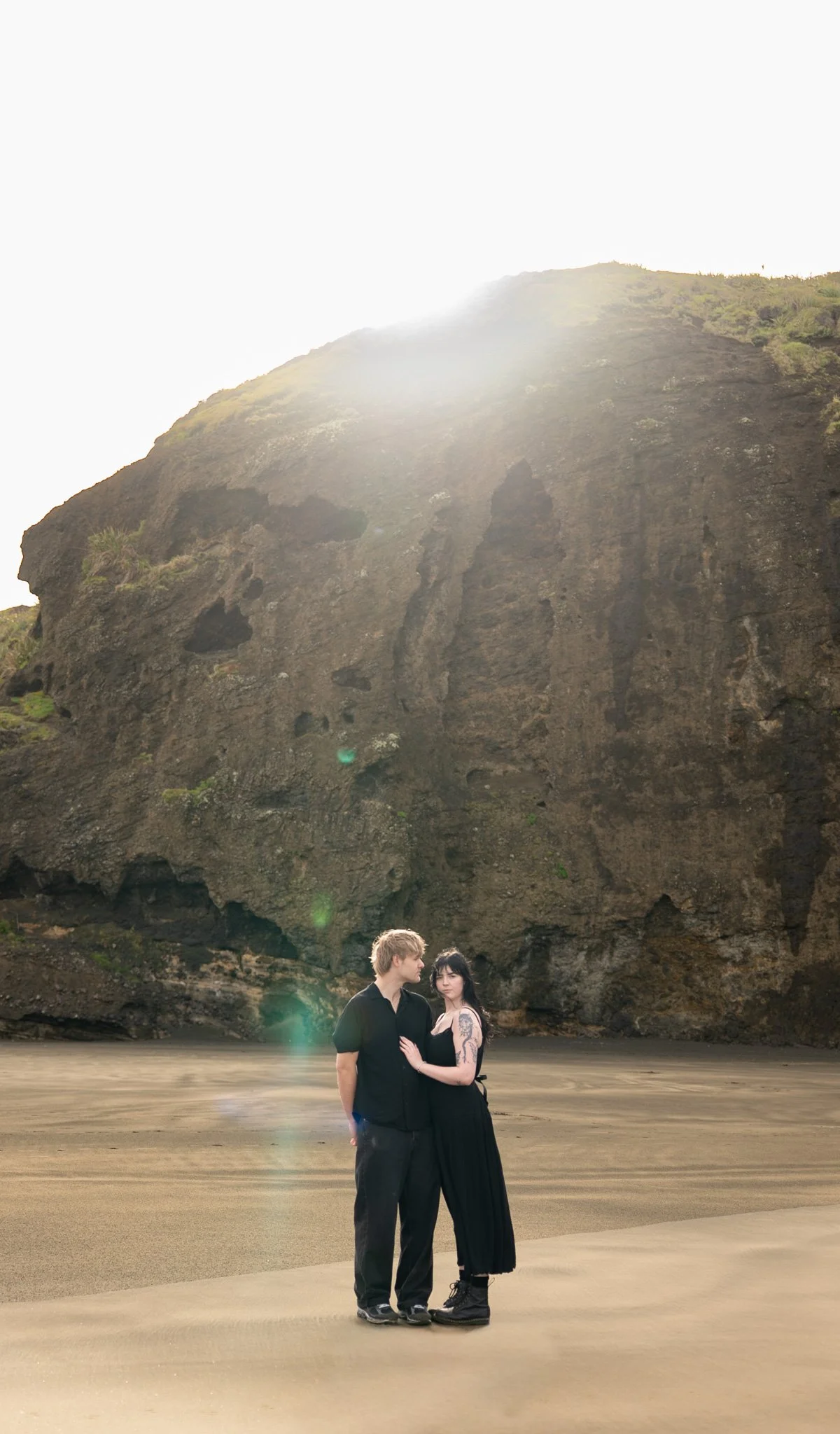 A couple standing on a sandy beach with a large rocky cliff in the background, sunlight shining over the top of the cliff.