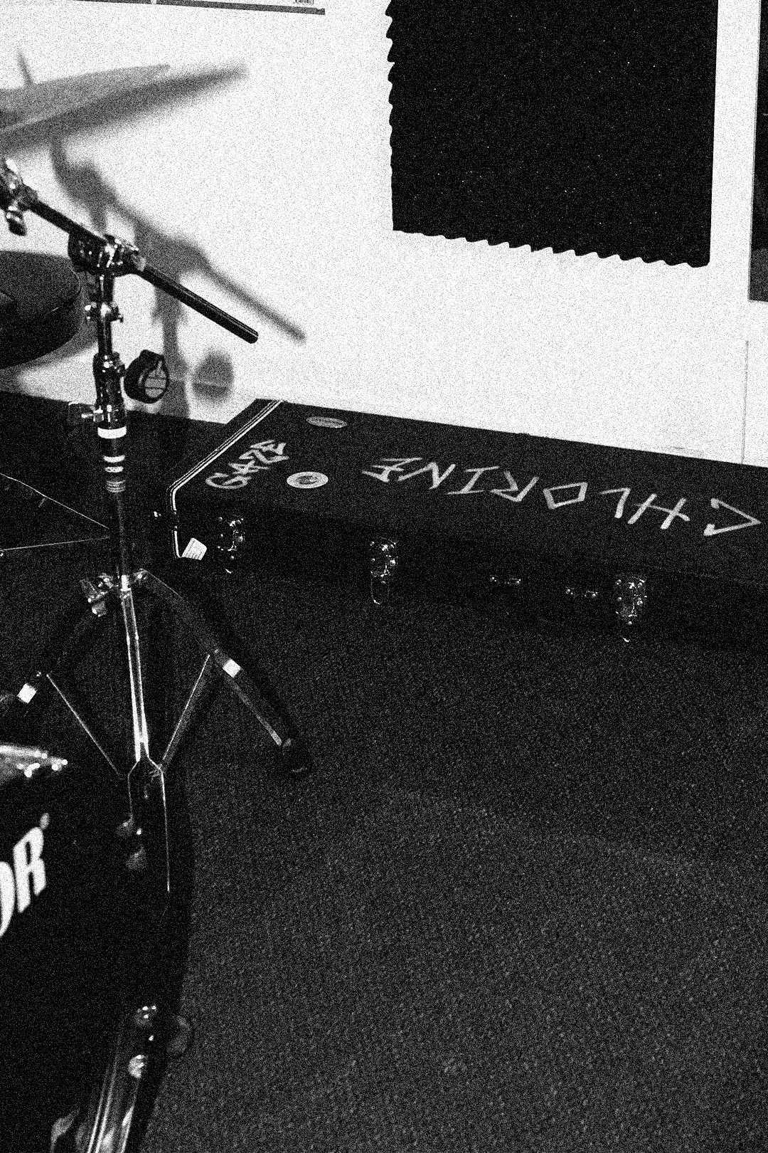 A black and white photo of a drum set on the left, with a bass drum and cymbal, and a black case labeled 'CHORD' on the floor in the background, along with a black foam soundproof panel on the wall.