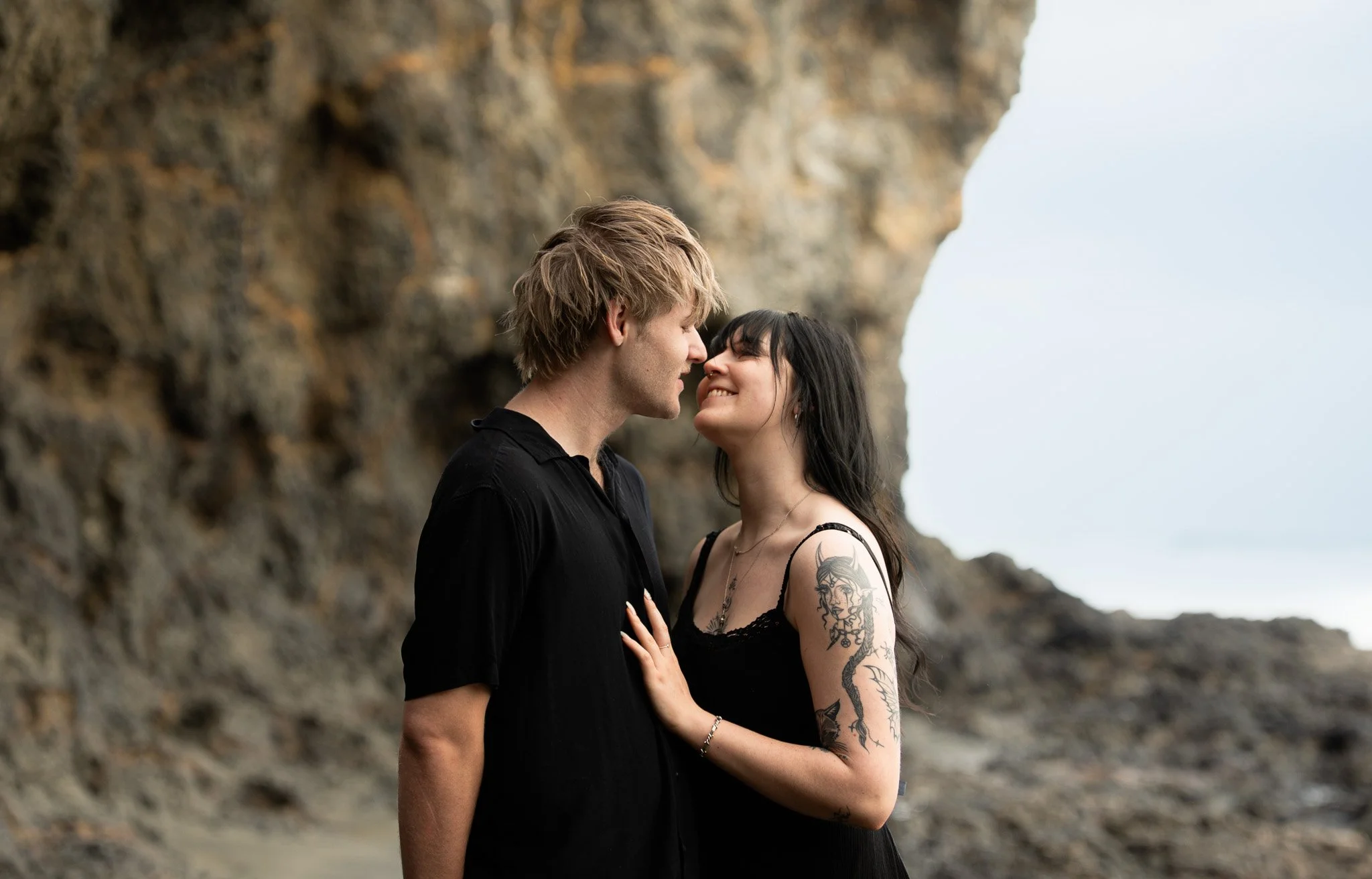 A young couple standing close together outdoors near a rocky cliff, facing each other with smiles, on a cloudy day.