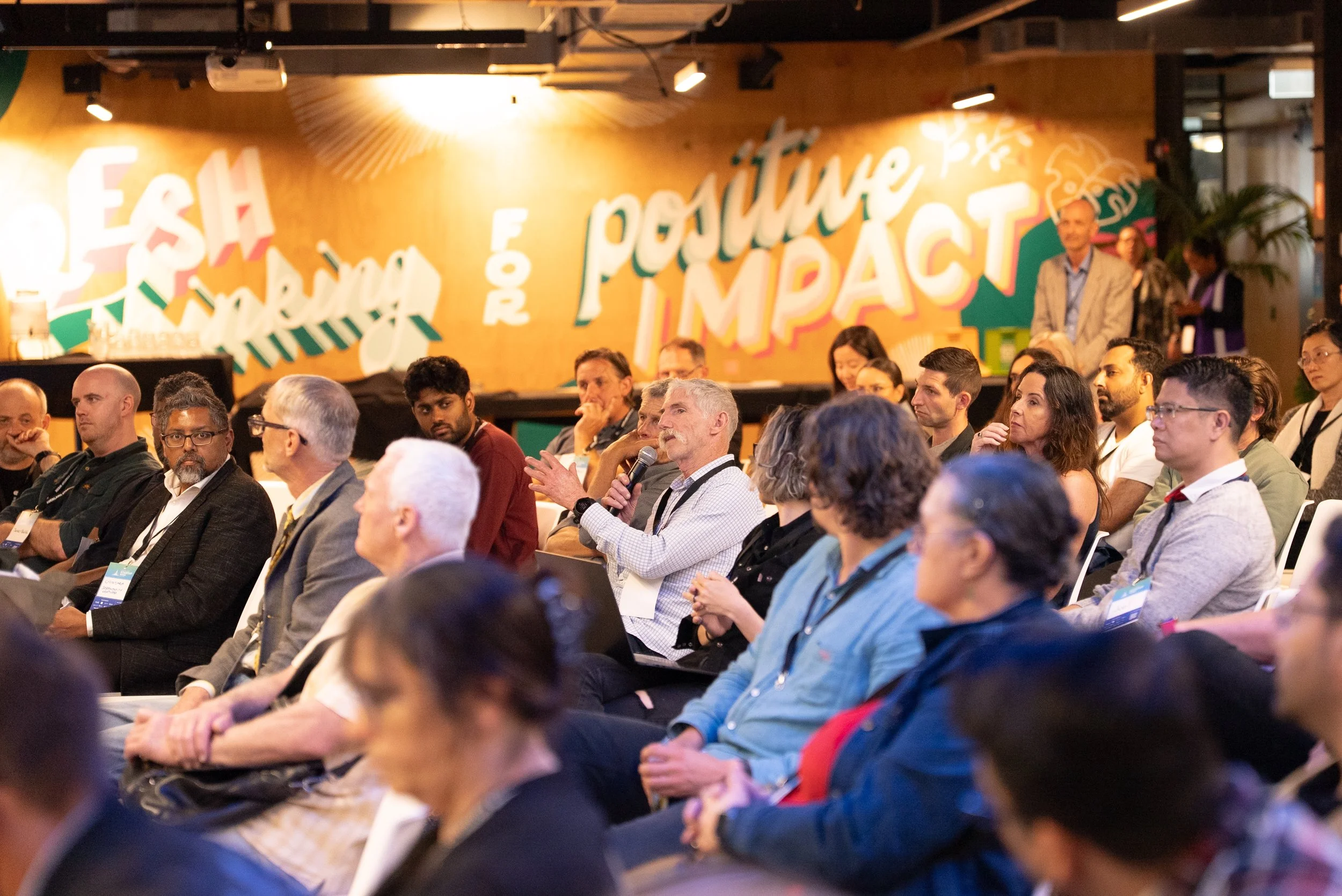 Audience members attentively listening at a conference with colorful wall art reading 'Fish for Change, Positively Impact' in the background.