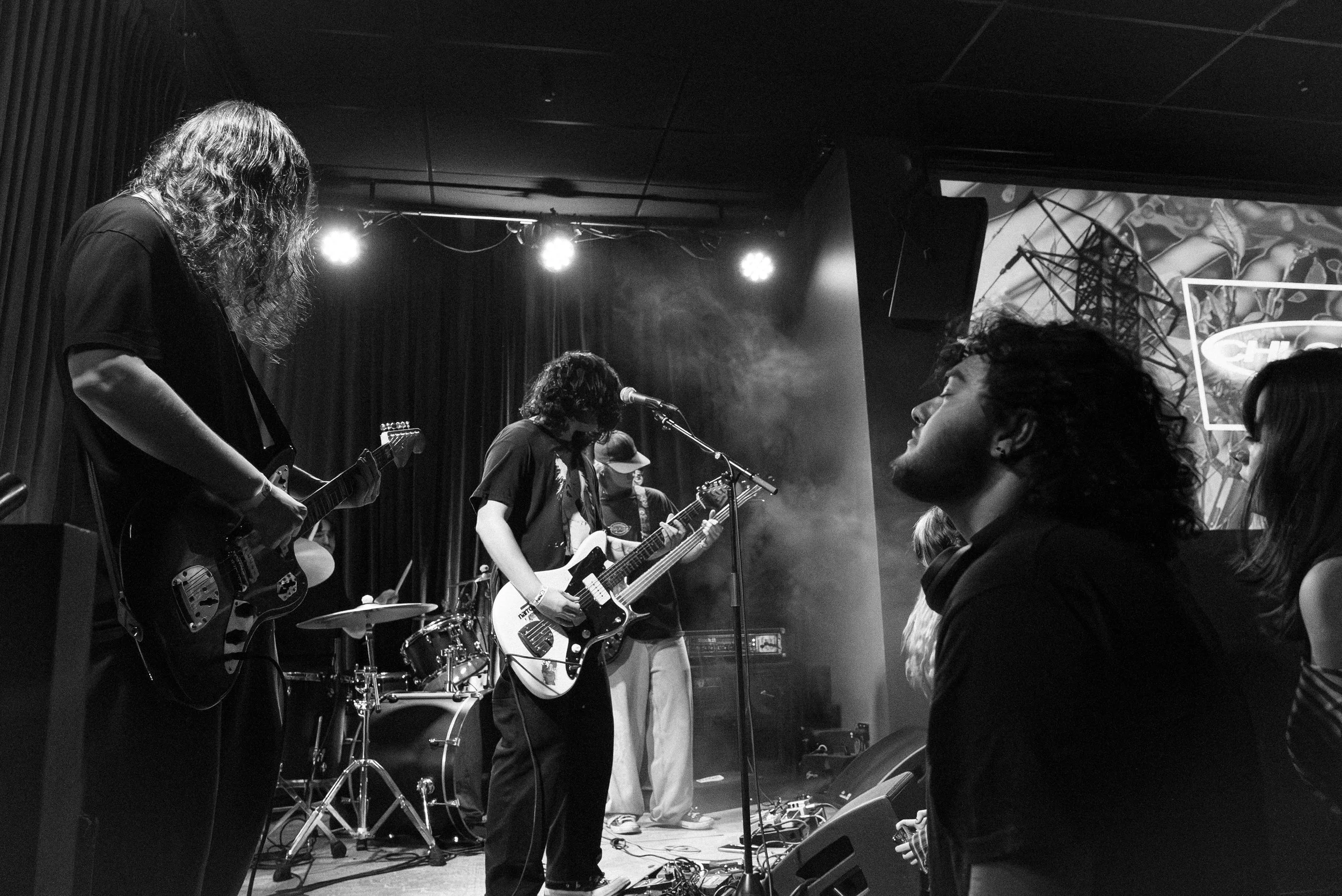 Black and white photo of a live music performance with band members playing electric guitars and a drummer on stage, and audience members watching.