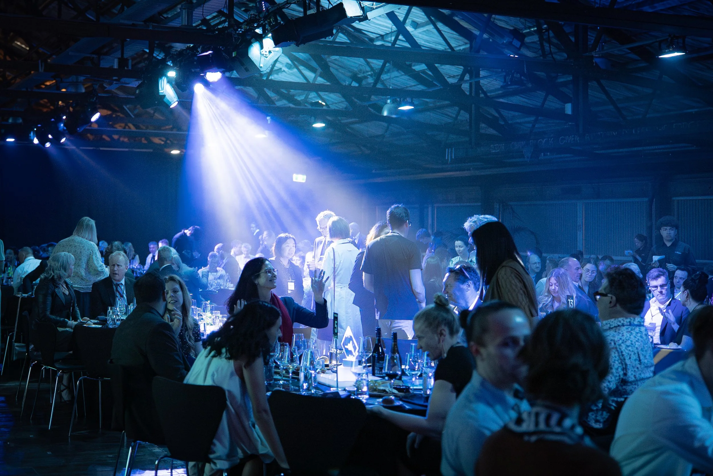 Crowd of people seated at tables and standing in a dimly lit event hall with blue lighting and a high ceiling.
