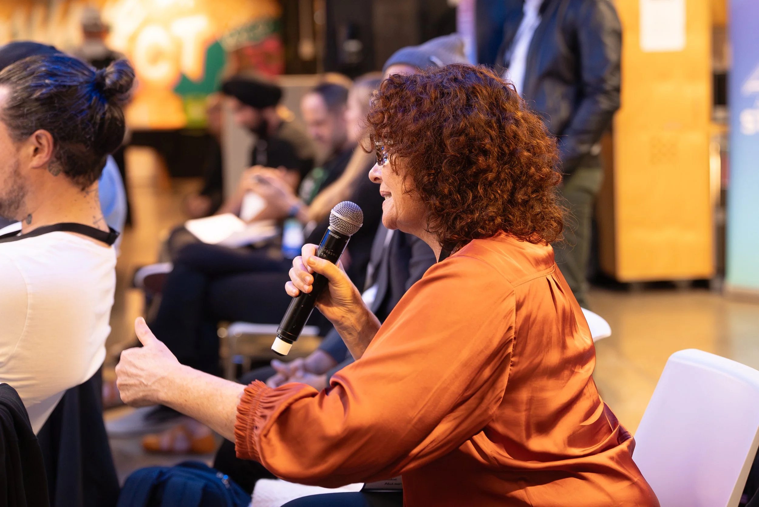 A woman with curly red hair and glasses holding a microphone, speaking at an indoor event. She is wearing an orange blouse and is engaged in a conversation with others seated nearby.