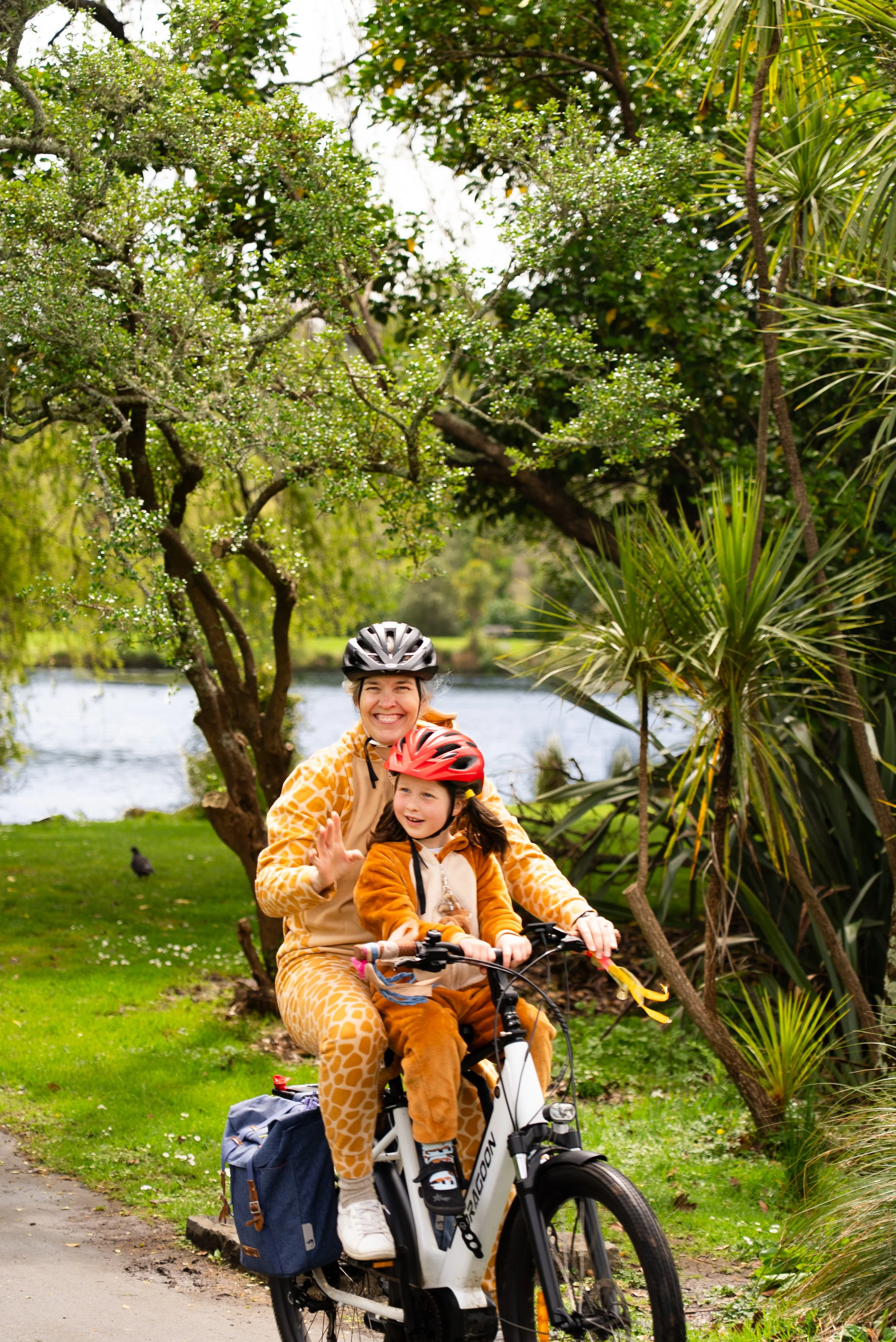 A woman and a young girl riding a bicycle outdoors near a lake, both wearing helmets and animal costumes, smiling and waving.