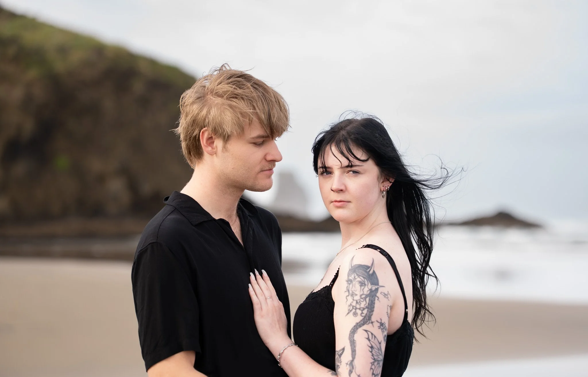 A young couple stands close on a beach with waves and cliffs in the background. The woman has black hair, tattoos, and is wearing a black tank top. The man has blond hair and is wearing a black shirt.
