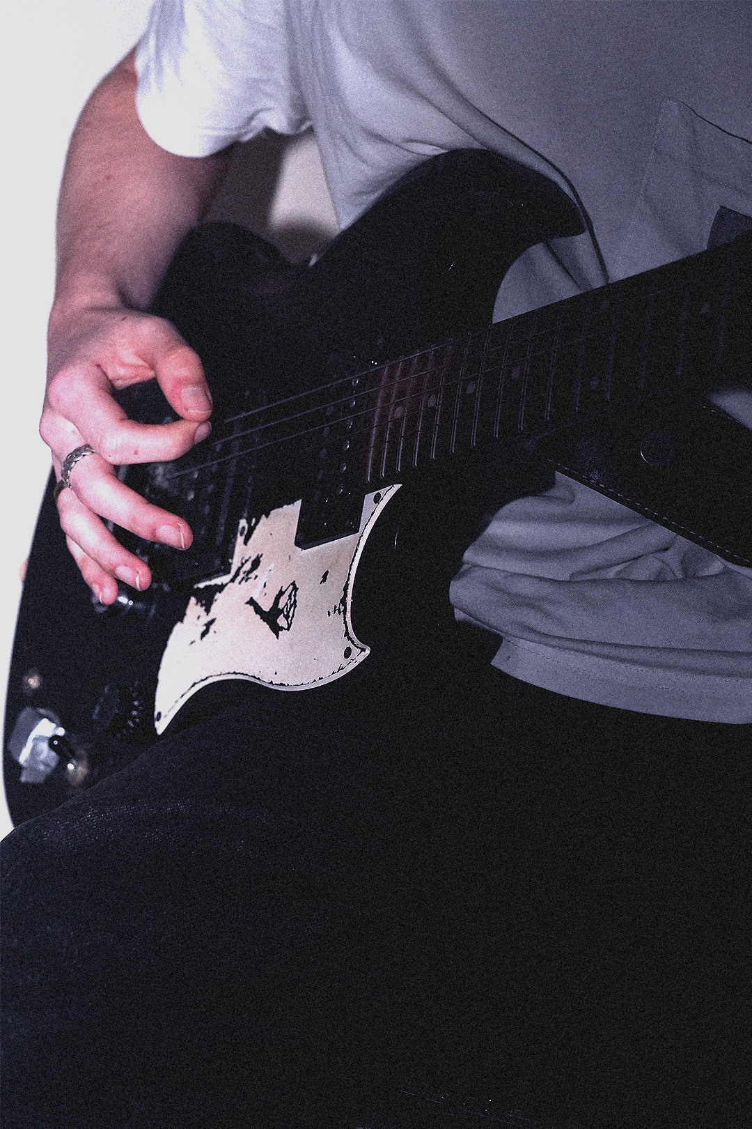 Close-up of a person playing a black electric guitar, wearing a white t-shirt and black pants.