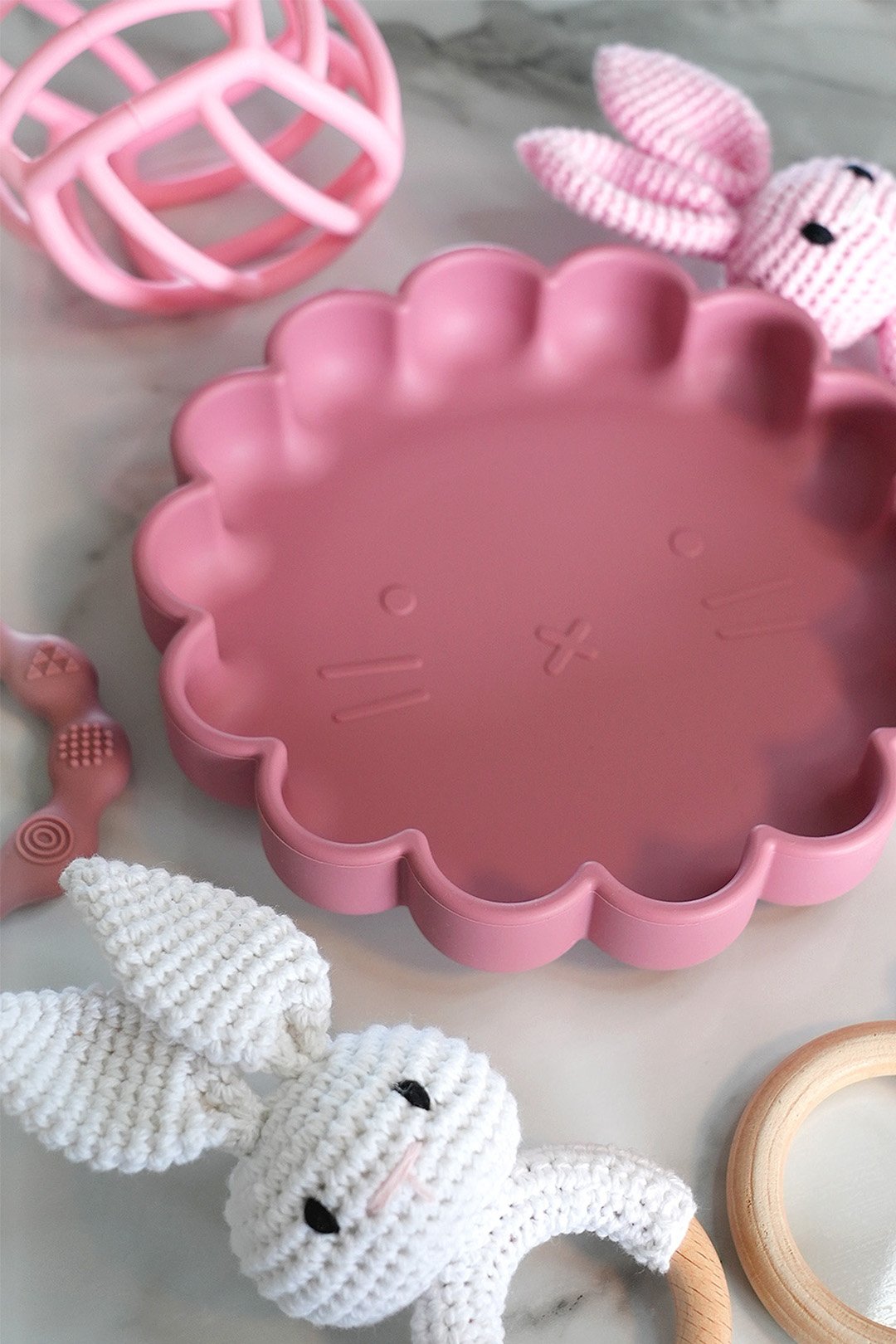 Assorted baby toys including a pink scalloped plastic plate, a white knitted bunny, a wooden ring, a pink plastic rattle, and a pink fabric bunny with a barcode pattern.