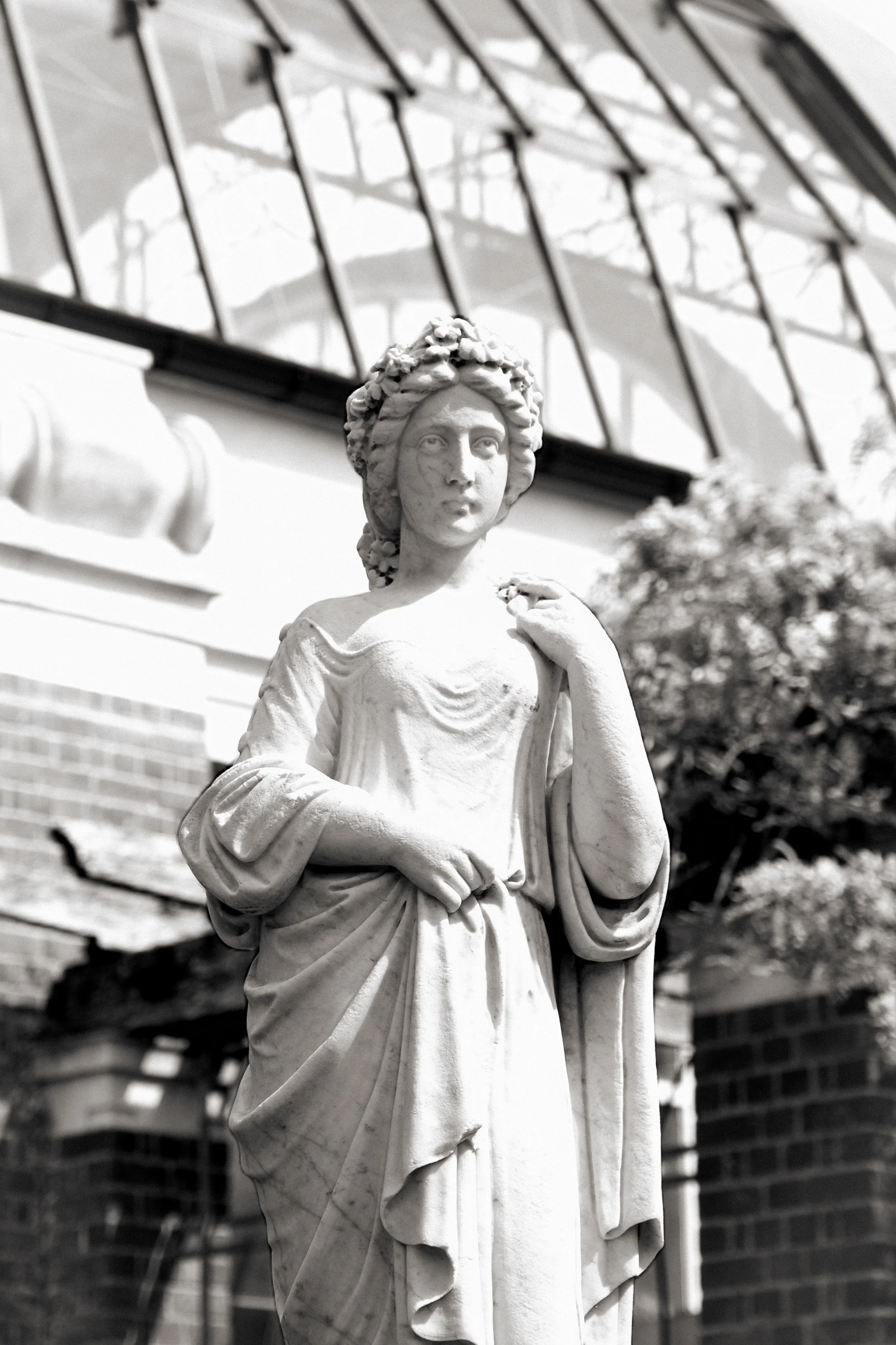 Black and white photo of a marble statue of a woman with a crown of flowers, standing outdoors in front of a building with large windows and trees.