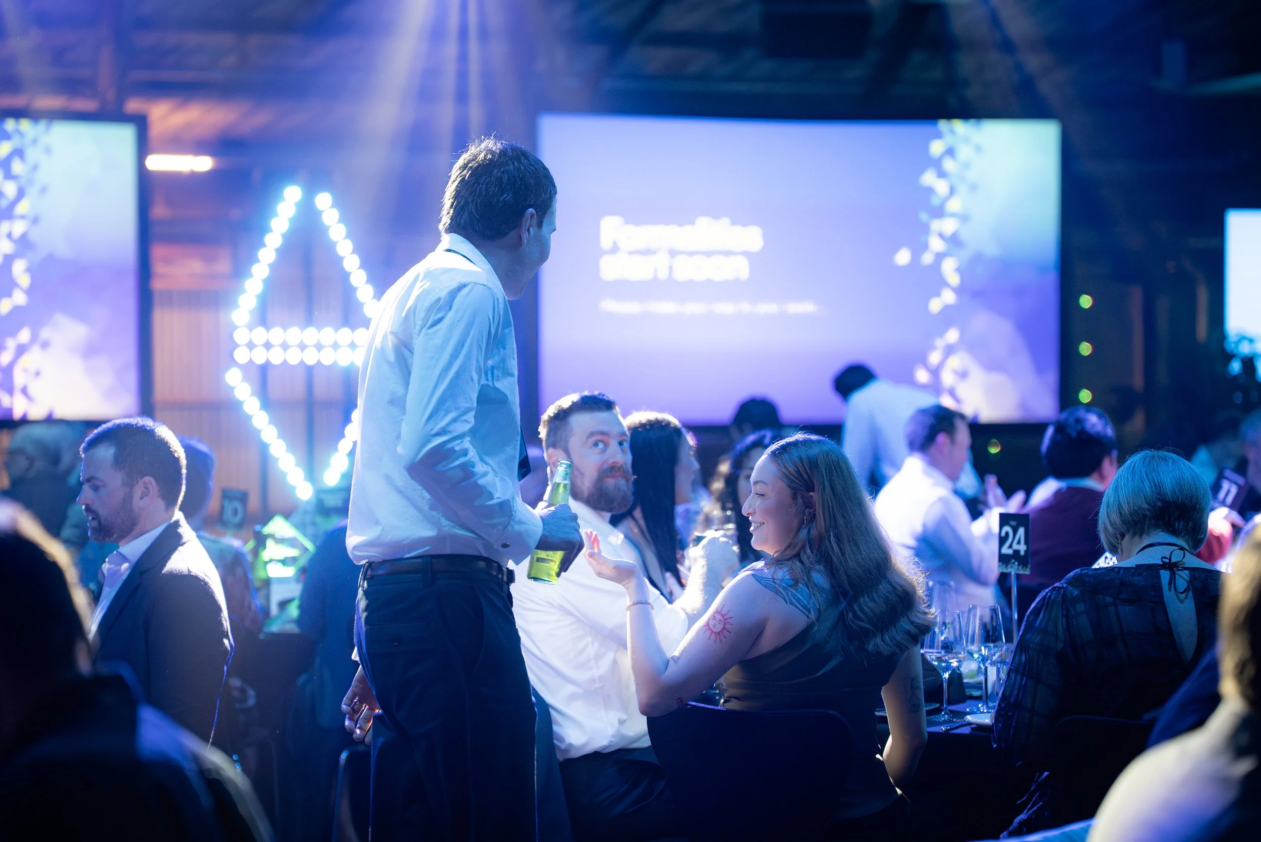 People attending a formal event or conference, seated at tables with wine glasses, in a well-lit venue with large screens and stage lighting.
