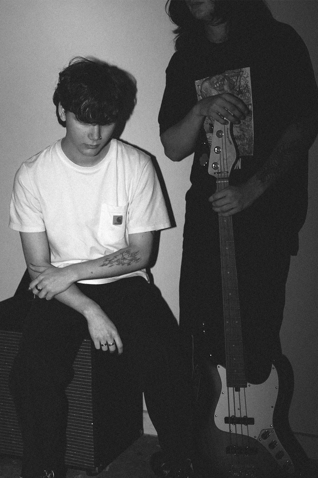 Black and white photo of two young men, one sitting on a stool or amplifier with arms crossed, the other standing holding a bass guitar, both with long hair and casual clothing.