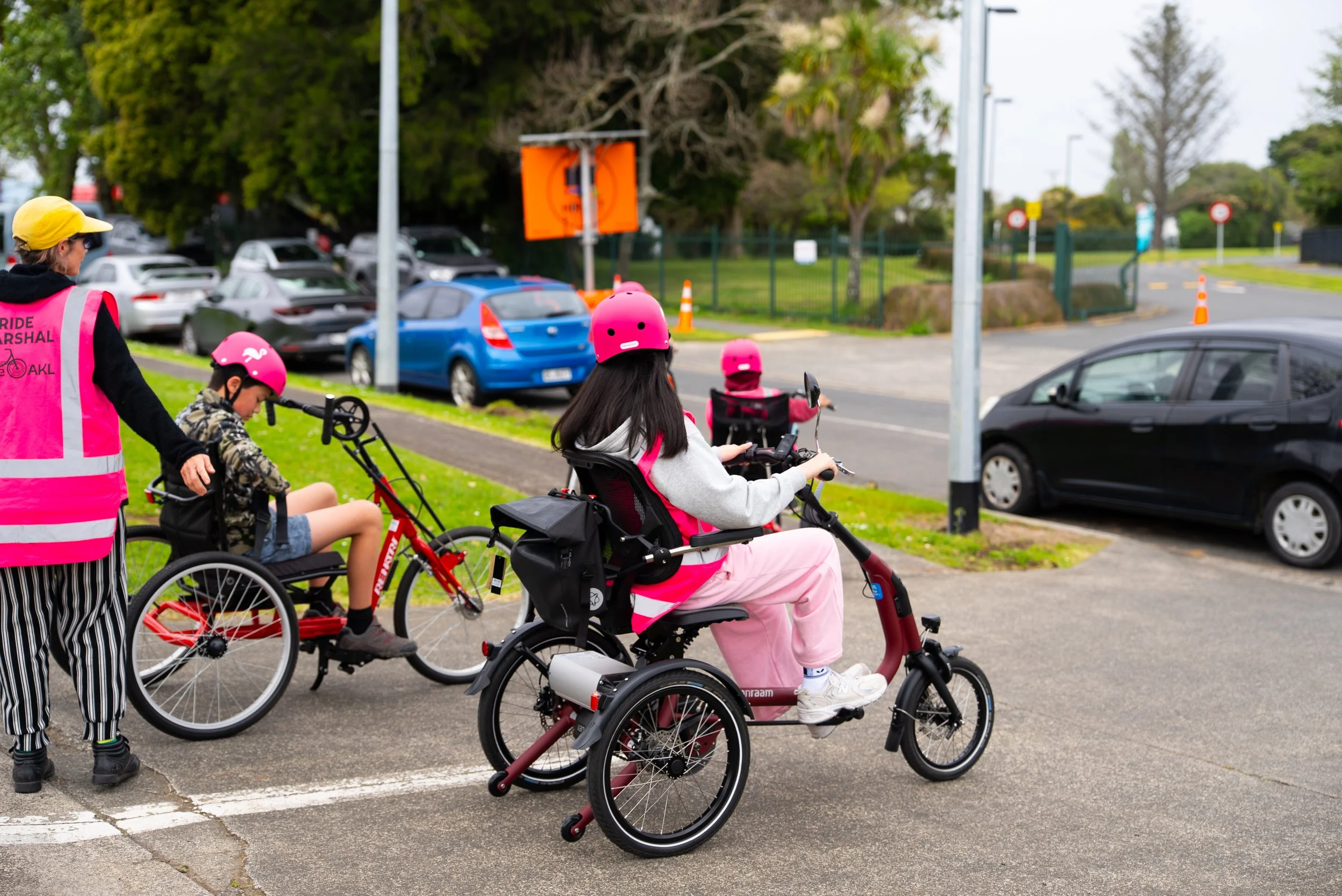 Children and an adult wearing pink helmets and vests, with one child using a tricycle and another in a wheelchair, preparing to ride bicycles together on a sidewalk in a park.