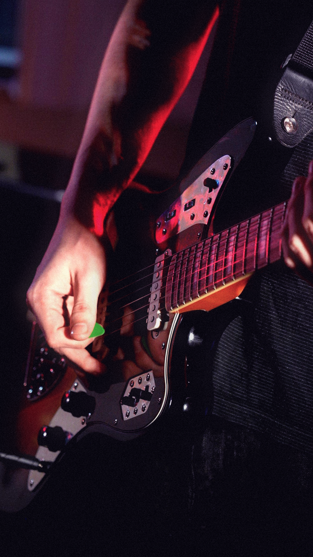Close-up of a person playing an electric guitar, with fingers on the strings and a red and black guitar body in a dimly lit setting.