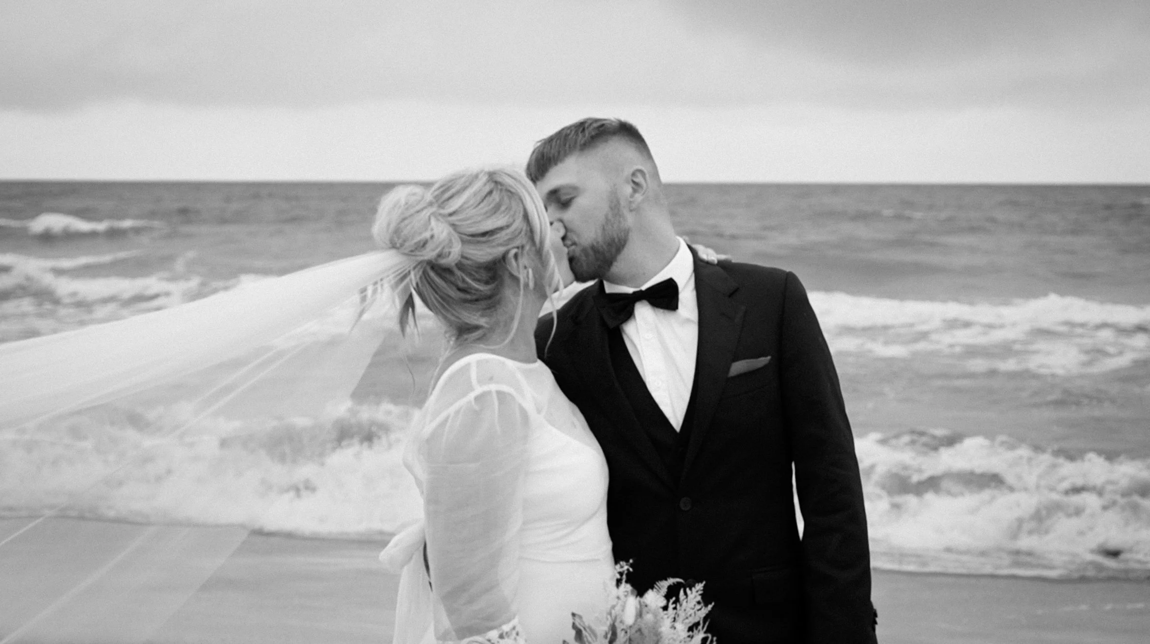 A black and white photo of a bride and groom sharing a kiss on the beach, with waves in the background.