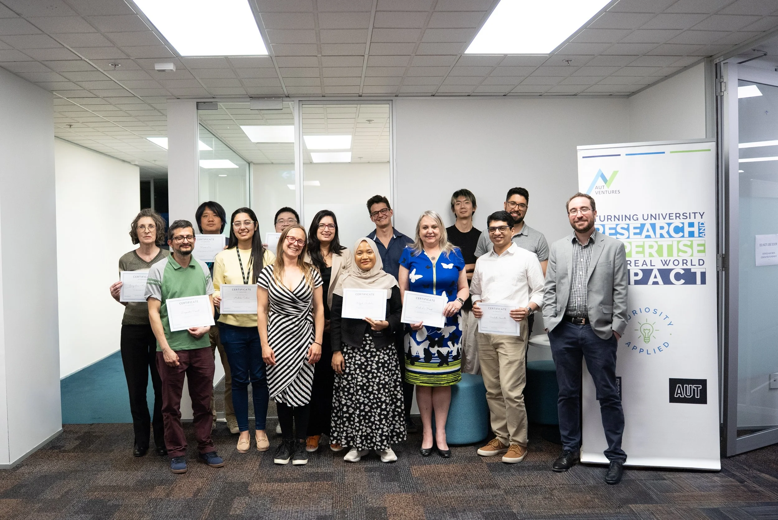 Group of diverse people holding certificates at an indoor event, standing in front of a banner that reads "Turning University Research into Expertise for a Real World Impact."