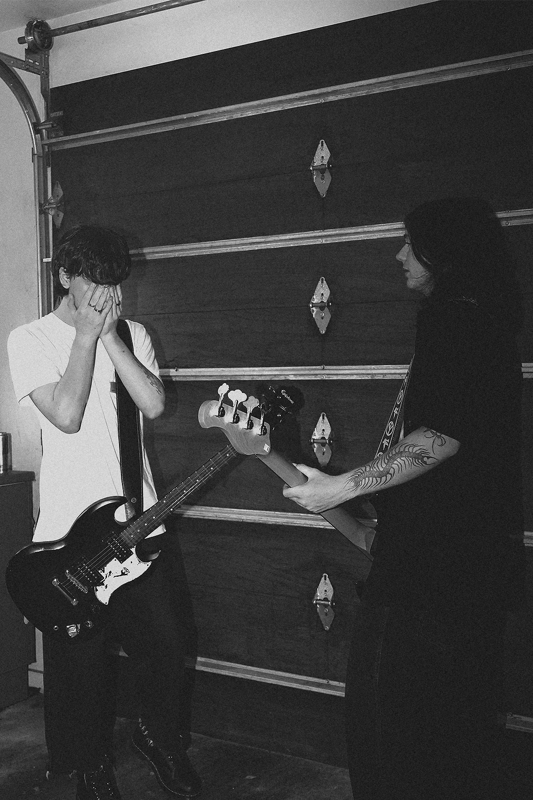 Two musicians, one with their face covered by their hands and the other with long hair and tattoos, are playing electric guitars in front of a garage door.