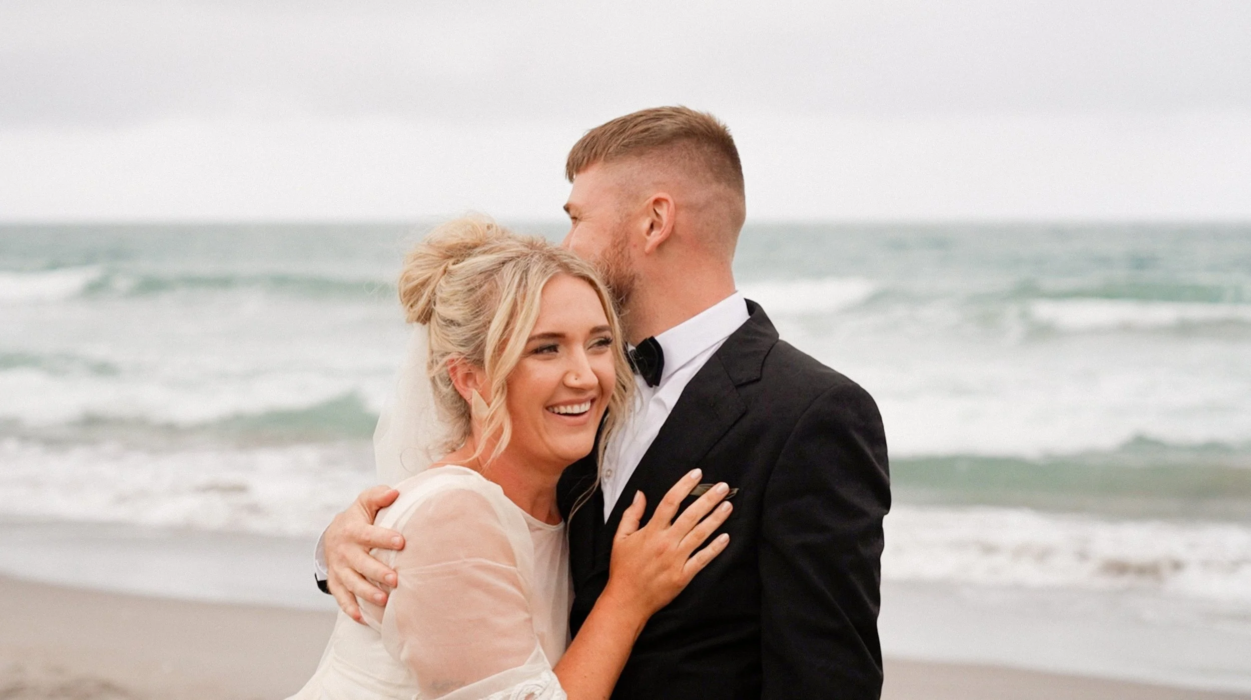 A bride and groom hugging on a beach, smiling, with ocean waves in the background.