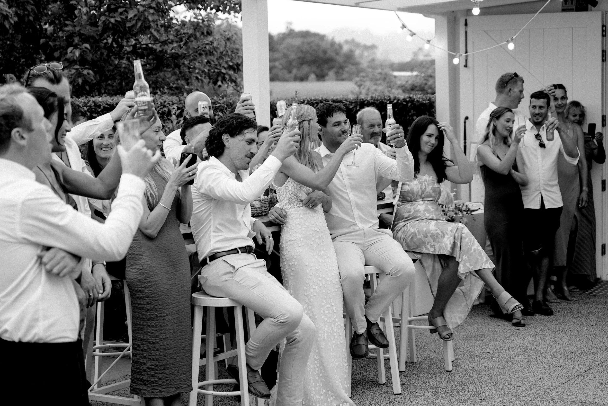 Group of people at a celebration, possibly a wedding, raising their glasses in a toast outdoors, some seated and some standing, with string lights overhead.