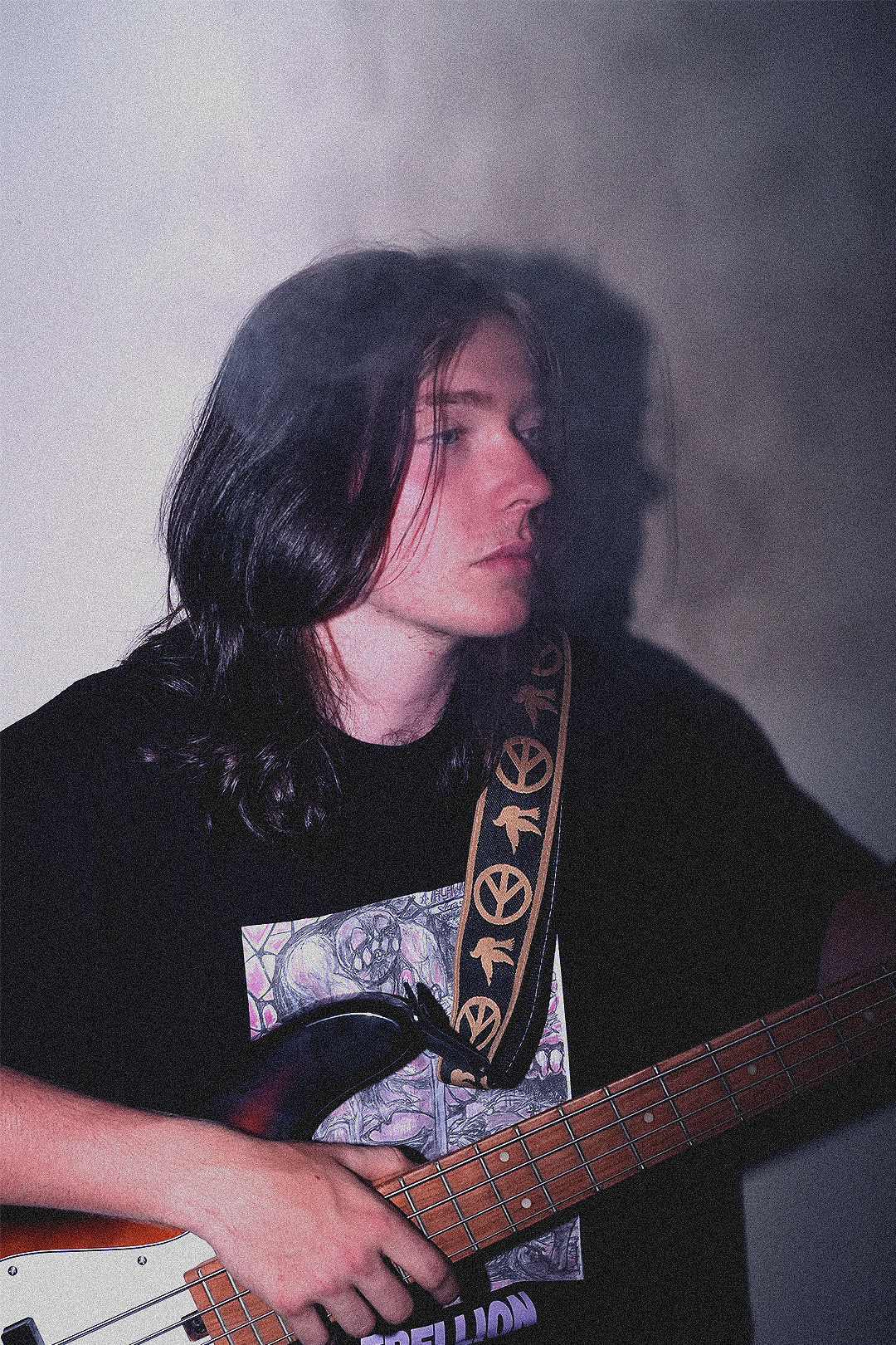 A young woman with long, dark, wavy hair playing an electric bass guitar. She is wearing a black T-shirt with a graphic print and a strap with peace symbols.