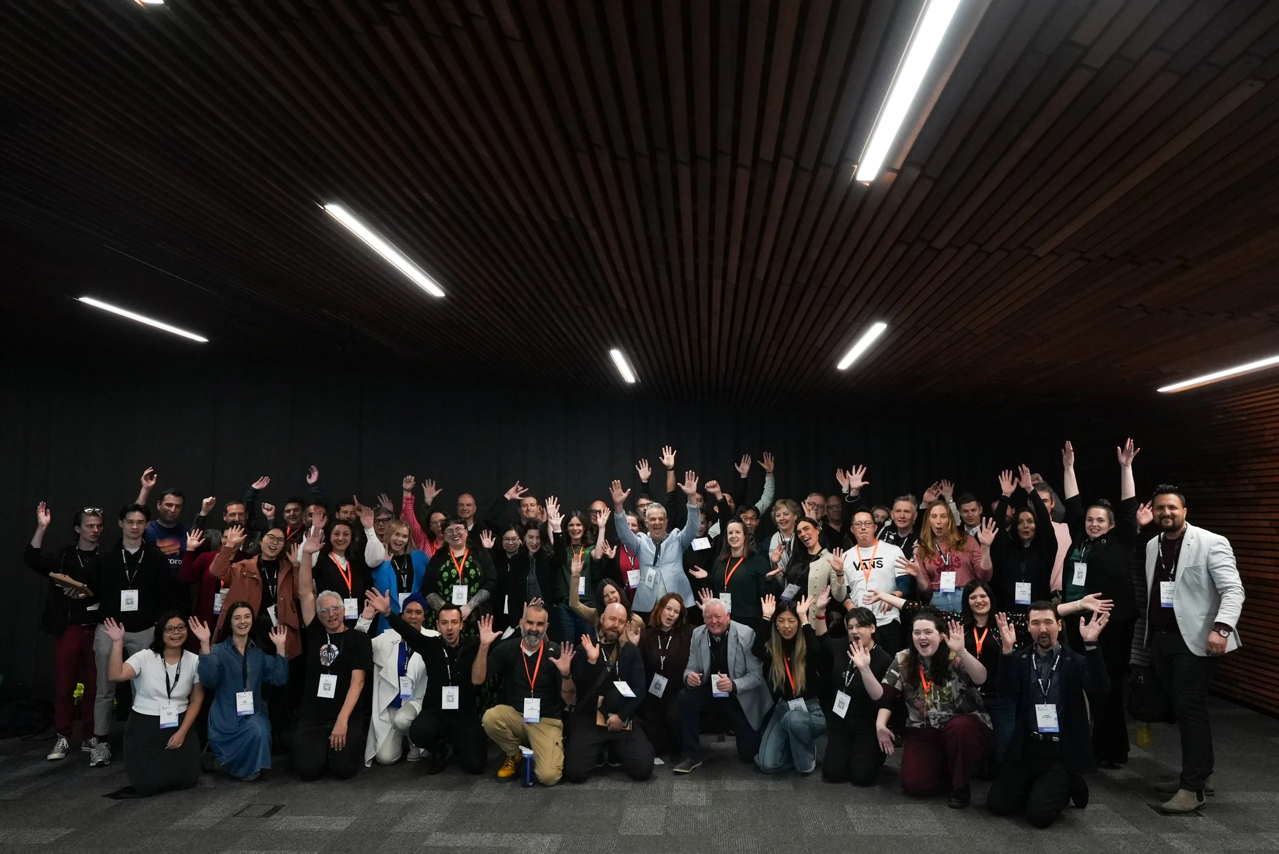 Group of diverse people at a conference or event, smiling and raising their hands, standing in a room with dark walls and wooden ceiling.